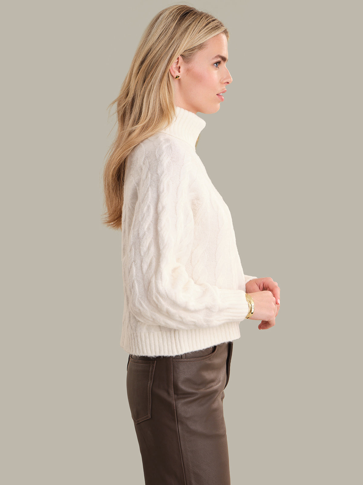 Woman wearing a white cable knit sweater and brown pants on a plain background