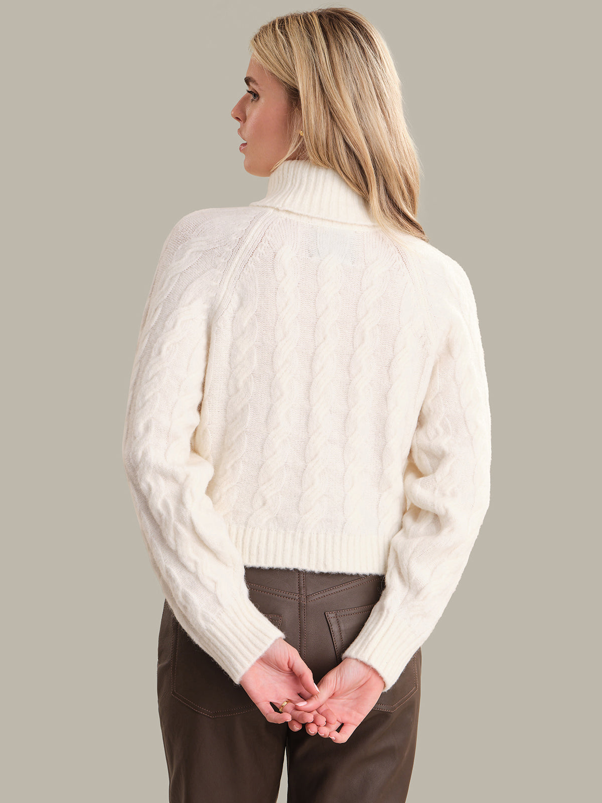 Woman wearing a white turtleneck sweater and brown pants on a plain background