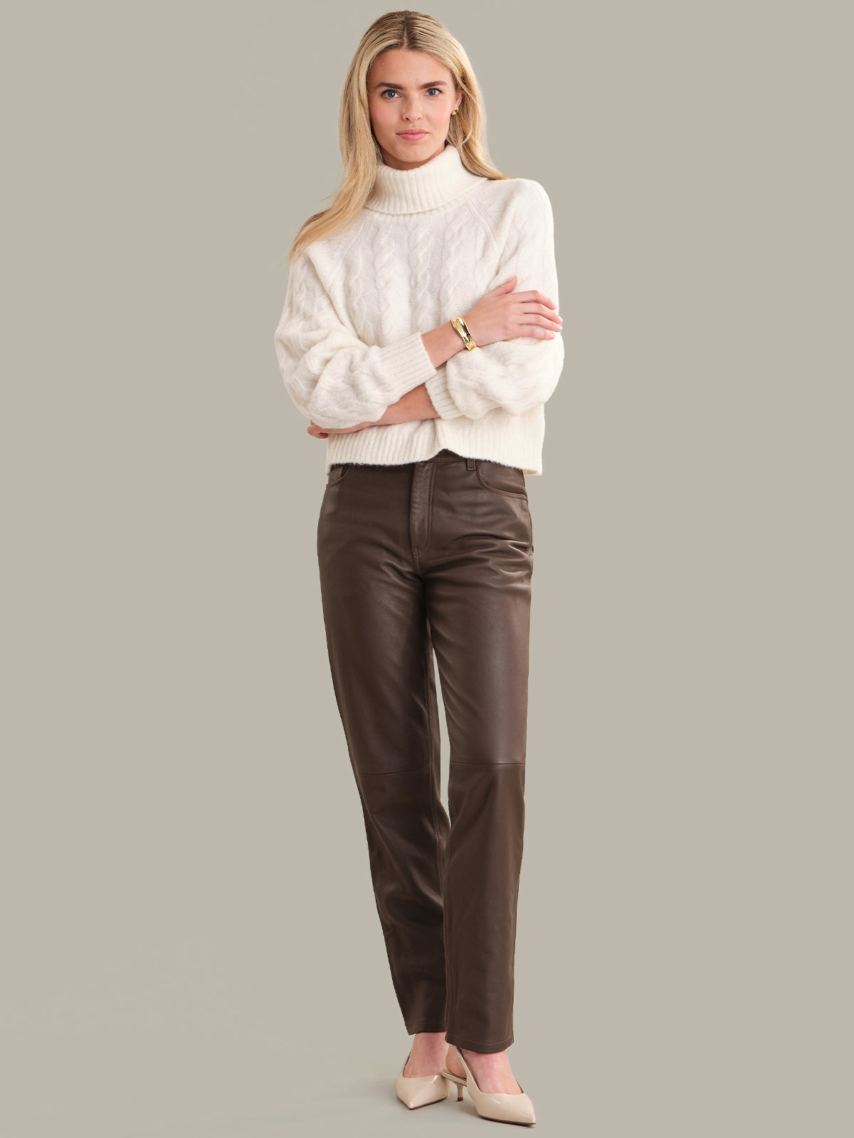 Woman wearing a white turtleneck sweater and brown leather pants on a gray background