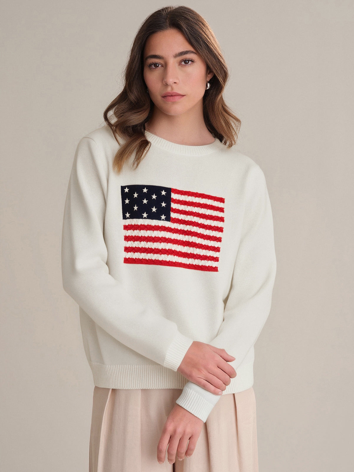 A woman with long brown hair models a 525 America 100% cotton crewneck sweater with a knit USA flag on the front. She stands before a beige background, gazing at the camera with a neutral expression.