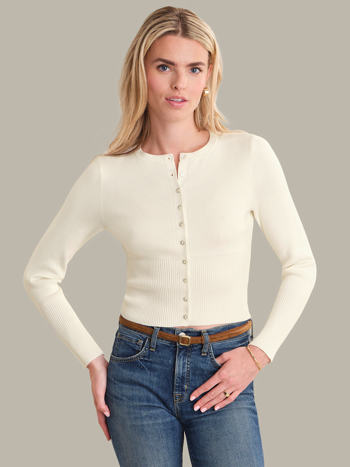 Woman wearing a cream cardigan over a black top with blue jeans on a gray background