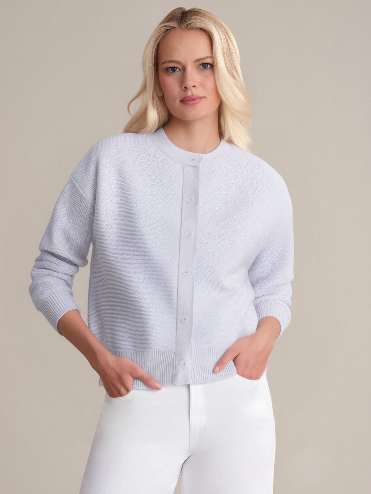 A person with long blonde hair wears a 525 America light gray button-up cardigan with a classic crew neckline and white pants, standing against a plain beige background with hands in pockets.