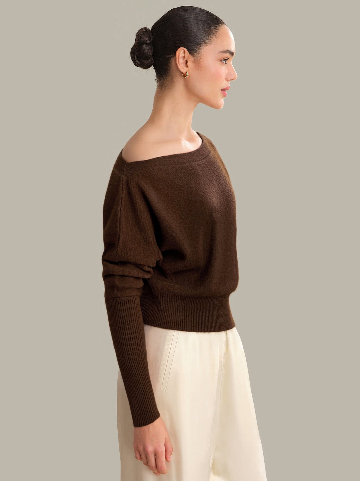 Woman wearing a brown sweater and white pants against a plain background
