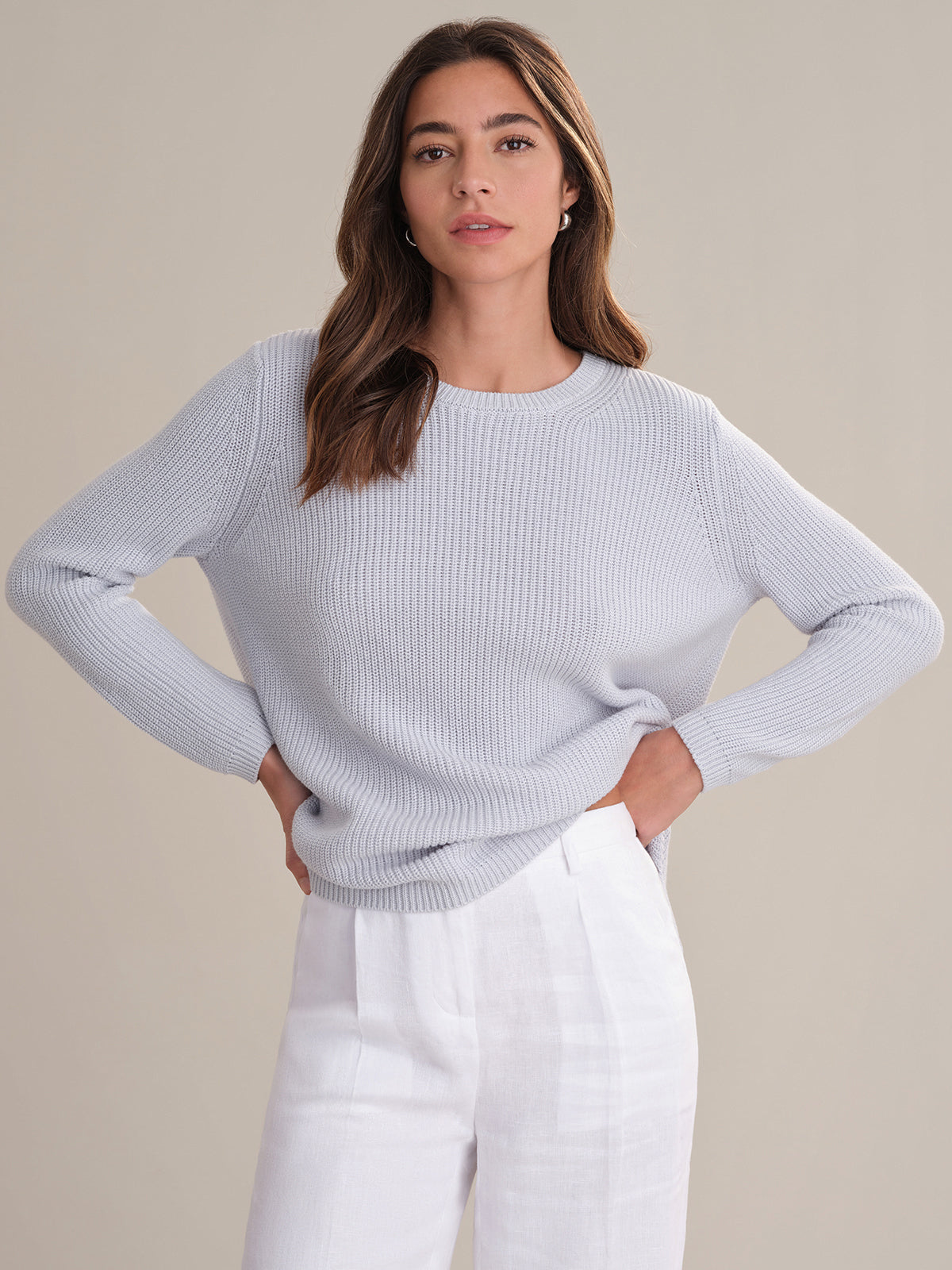 A woman with long brown hair wears a 525 America light grey shaker stitch knit sweater and white high-waisted pants, standing against a beige background with her hands on her hips.