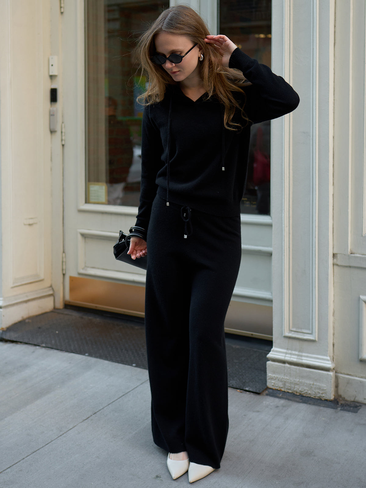 Dressed in luxury, a person stands by an ornate building entrance, wearing sunglasses and an all-black ensemble featuring Willow Cashmere Straight Leg Pants, a matching sweater, chic white pointed shoes, and stylishly holding a black handbag.