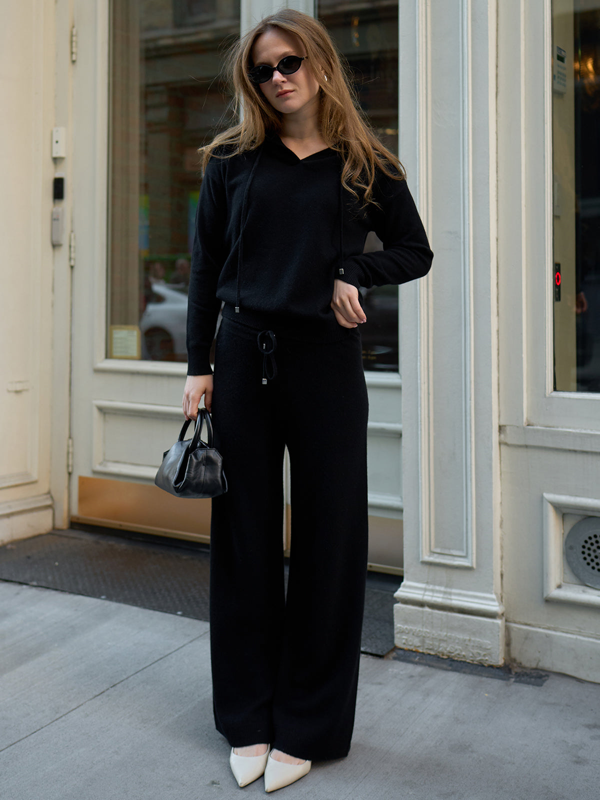 A person with long hair, wearing sunglasses and a black outfit, stands outside a building with white-trimmed doors and windows. Exuding luxury fashion, they hold a small black handbag and wear white pointed shoes paired with chic Willow: Cashmere Straight Leg Pants.