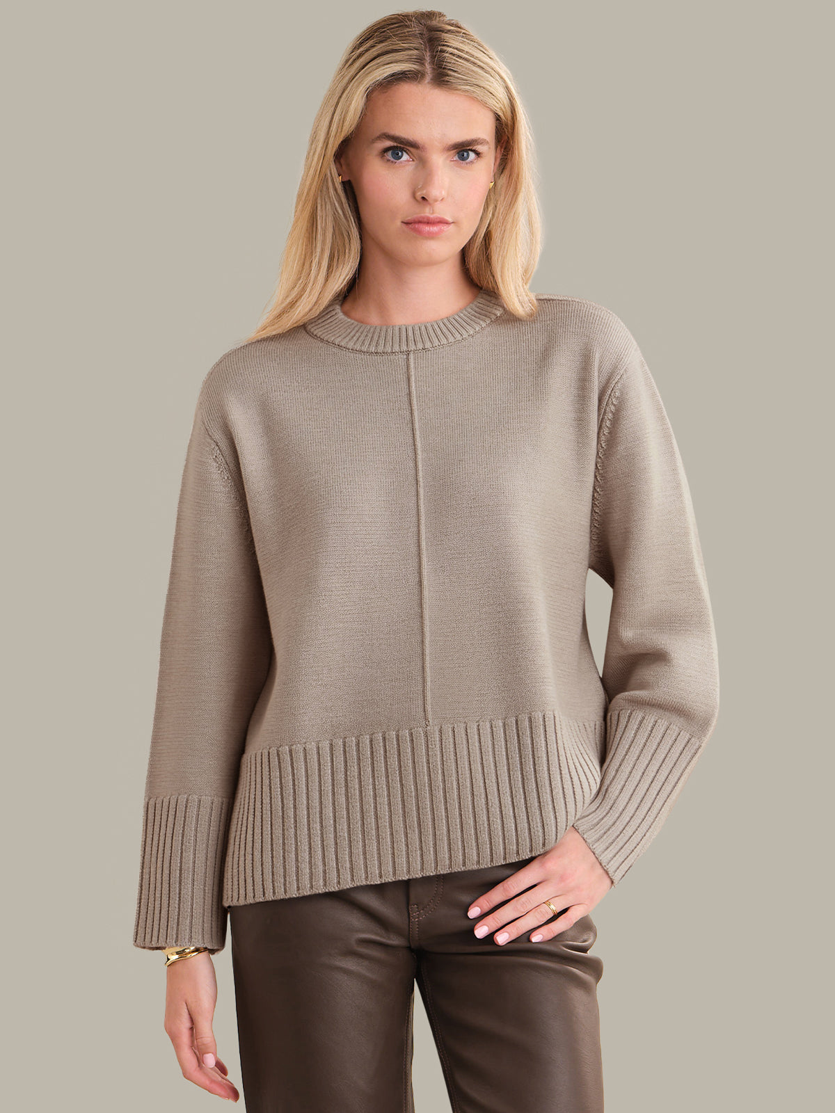 Woman wearing a beige sweater and brown pants against a plain background