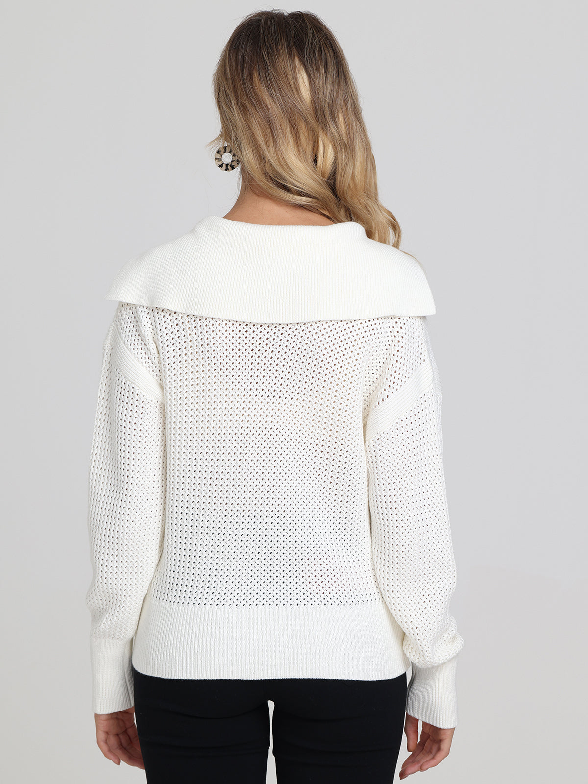 A woman with long, wavy blonde hair is shown from the back wearing a 525 America white knit pullover sweater with a wide collar, textured open-knit pattern, relaxed fit on the back and sleeves, paired with black pants.