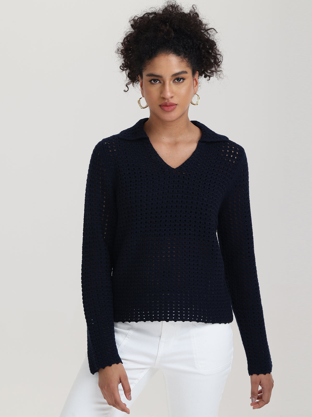 A person with curly hair stylishly dons a dark blue, relaxed-fit V-neck knit sweater featuring a collar from 525 America. They effortlessly pair it with white pants and hoop earrings, standing against a simple gray background.