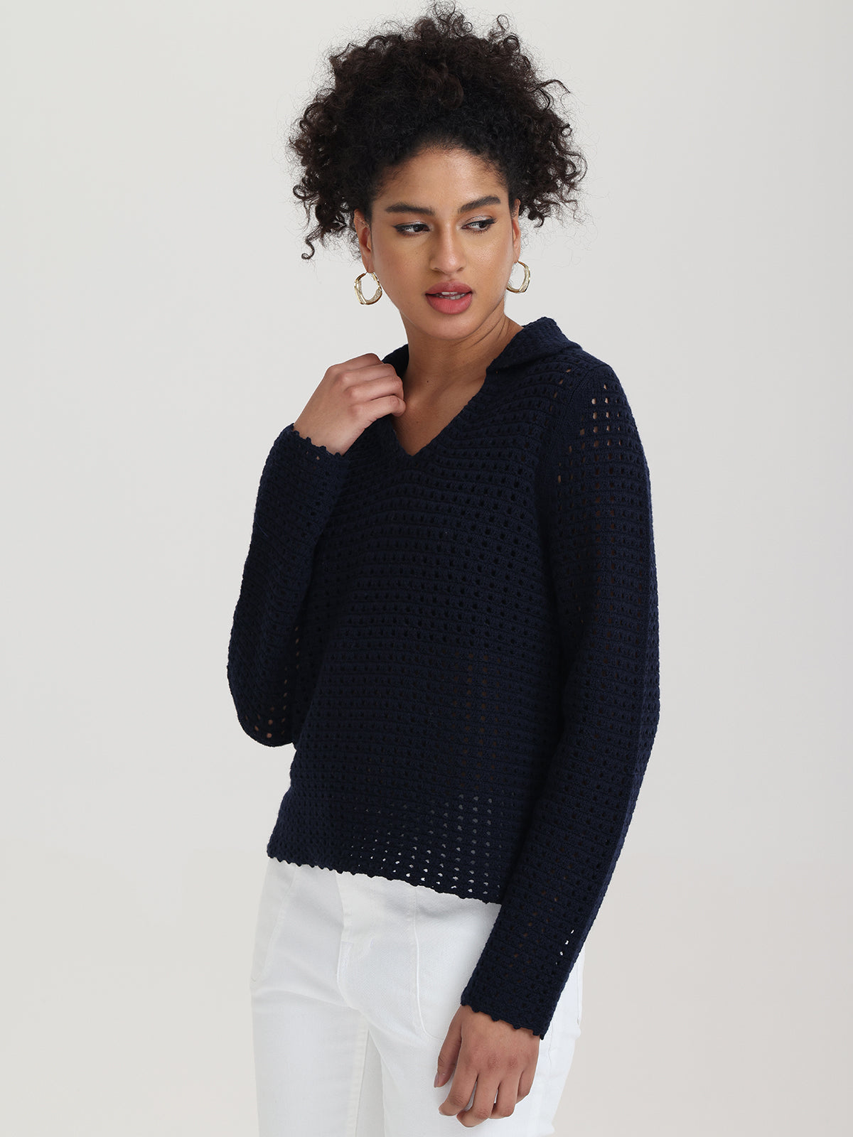 Wearing a 525 America navy blue openwork V-neck sweater with long sleeves and white pants, the person, with curly hair cascading, gently touches their neck while glancing sideways against a light backdrop.
