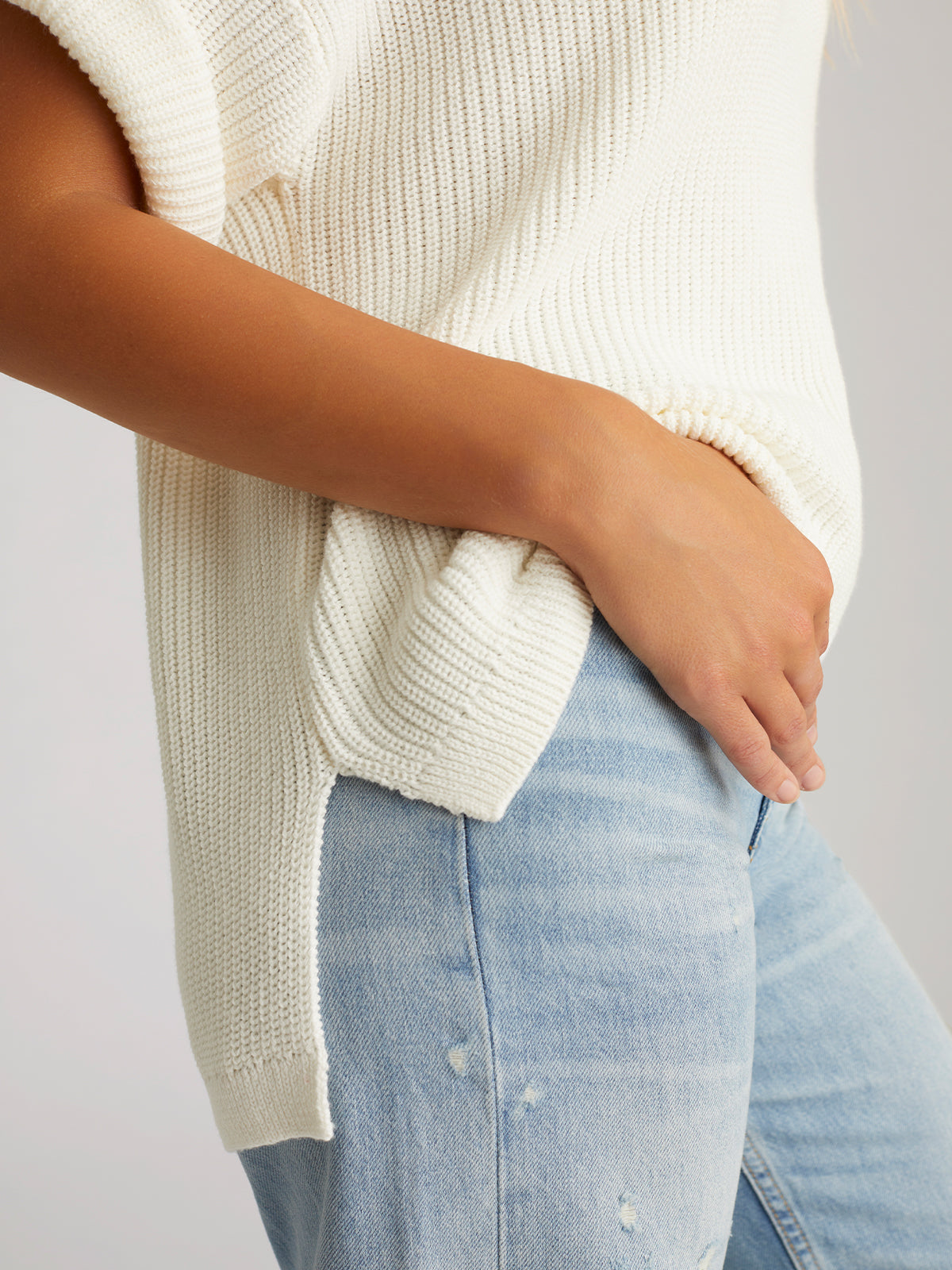 Wearing a cream Emma Crewneck Shaker Stitch Tee, made from 100% cotton with a side slit, paired with faded blue jeans featuring subtle distressing. The persons hand rests casually in a pocket against a light background for timeless comfort.