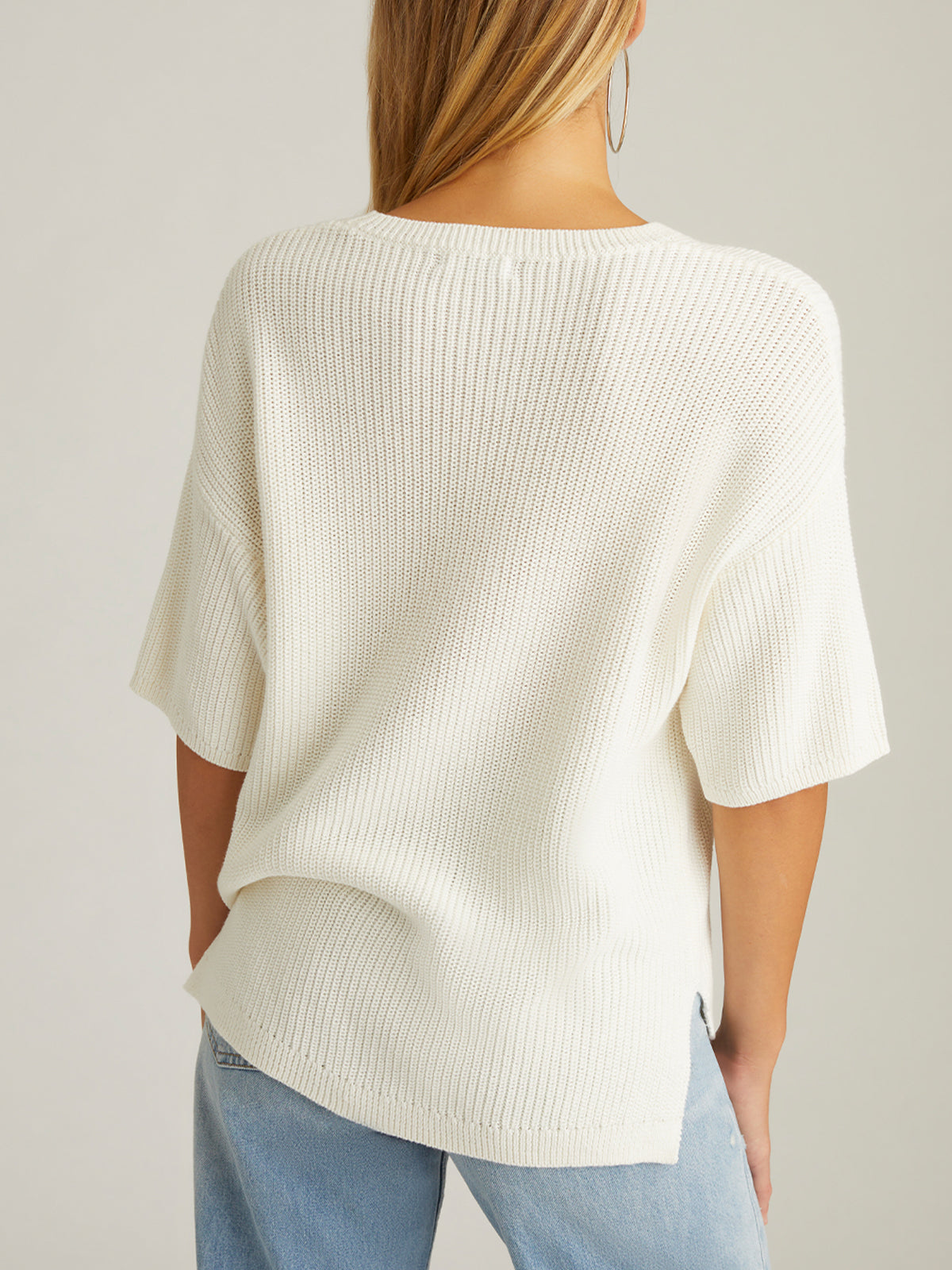 A person with long hair, facing away, is in a white Emma Crewneck Shaker Stitch Tee and light blue jeans. The tee, made of 100% cotton with a ribbed texture and short sleeves, stands out against the plain neutral background.