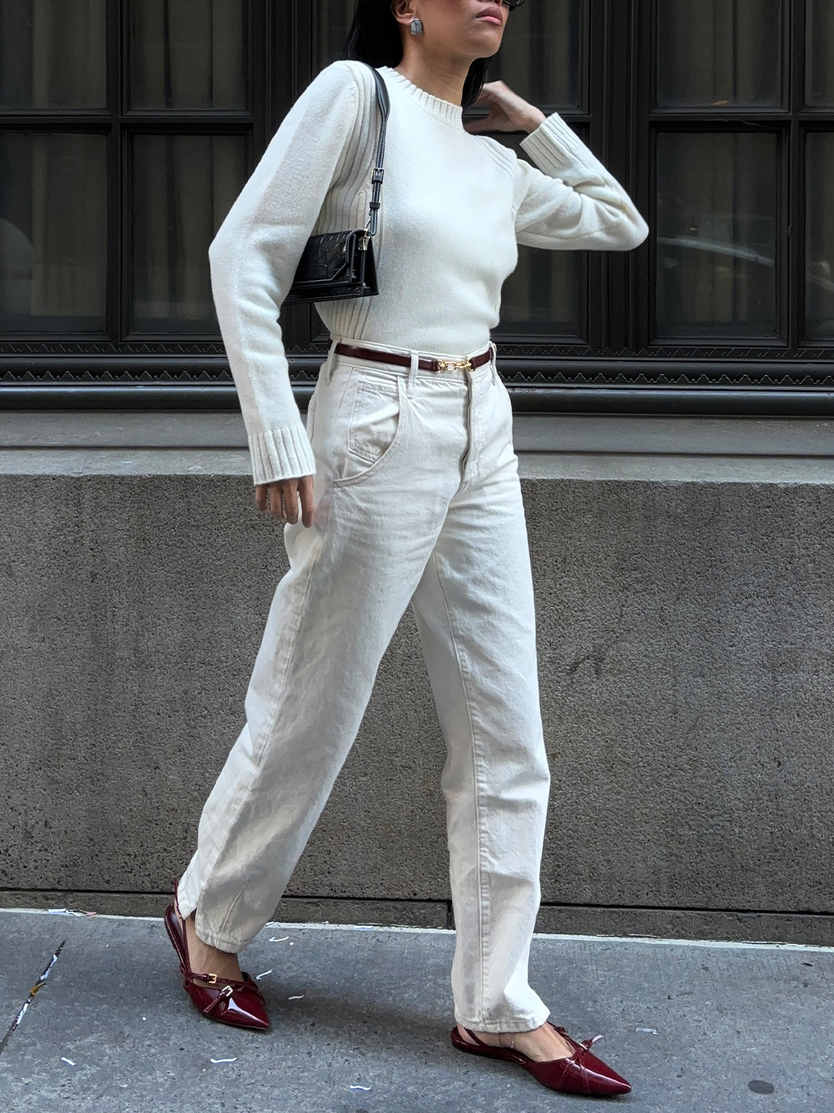 As a person walks along the sidewalk, they wear a white 525 America cashmere crewneck sweater and cream jeans, paired with a small black shoulder bag and red pointed shoes. The scene is enhanced by large, dark-framed windows and a stone wall in the background.