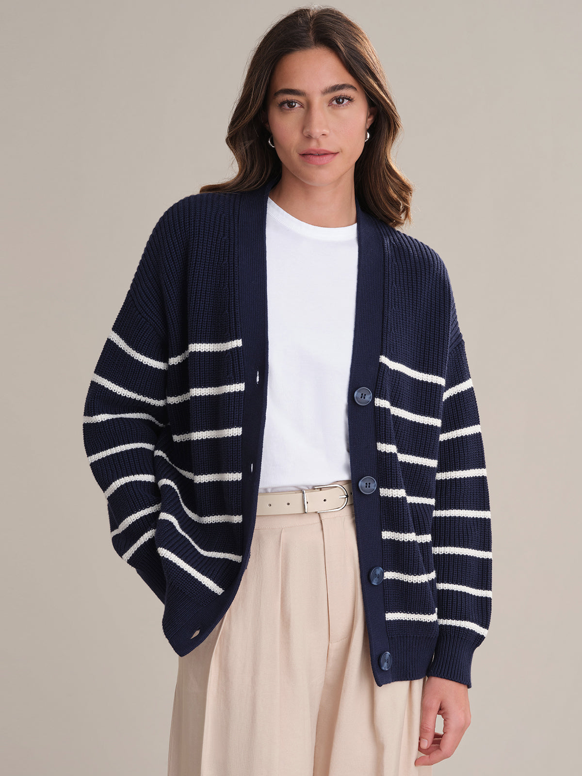 A woman with long brown hair wears a 525 America oversized navy blue cardigan with white stripes over a white tee and beige high-waisted pants, embracing a relaxed fit as she stands against a plain light gray background.