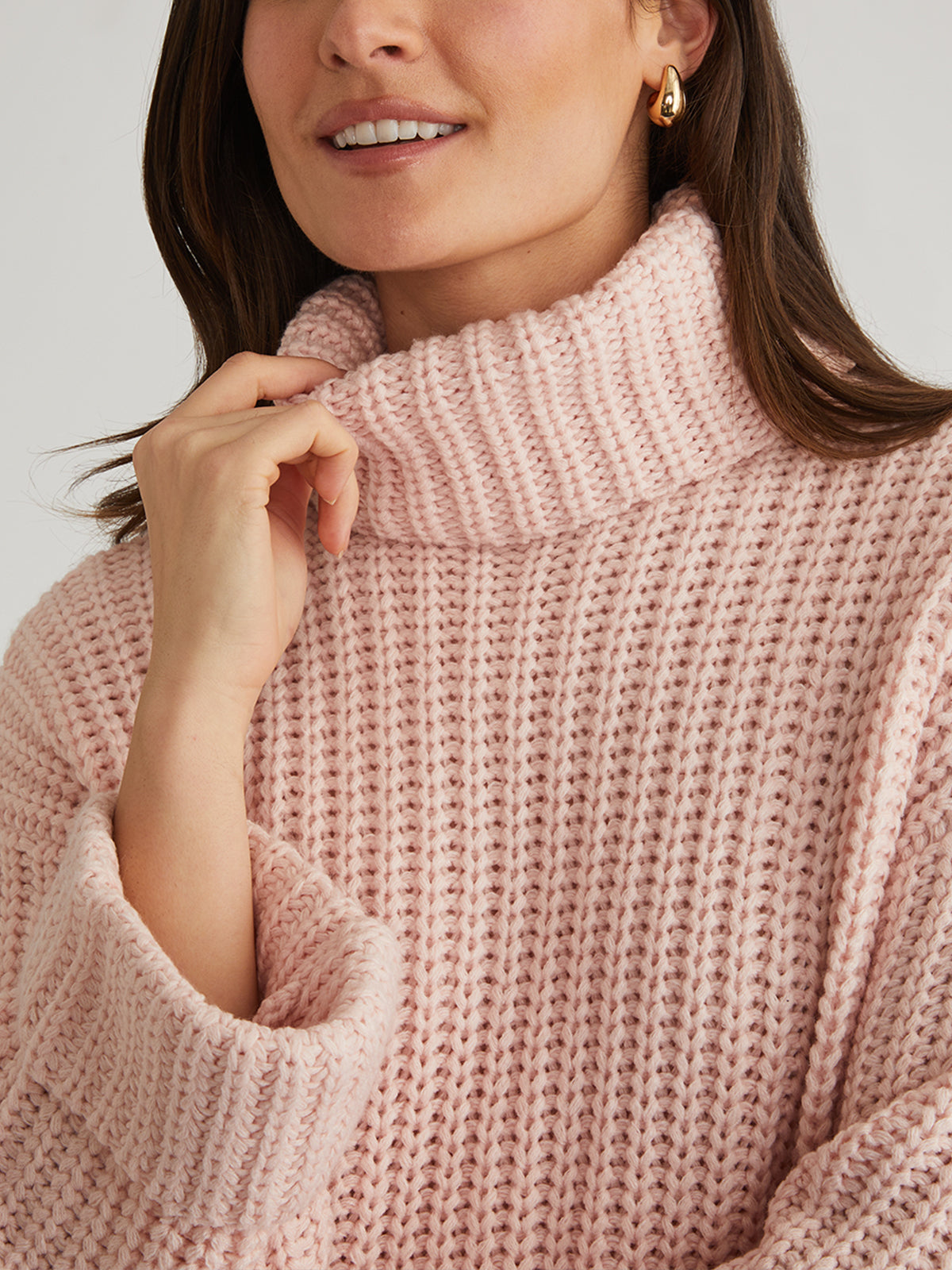 A woman wears a pale pink chunky knit 525 America Turtleneck Shaker Pullover with a cozy turtleneck design and gold hoop earrings, holding the collar with one hand. The image is cropped to show her from the shoulders up.