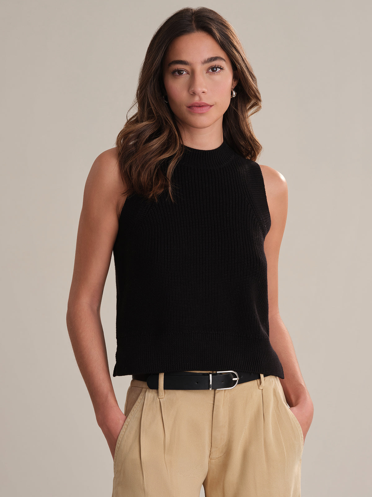 A person with long brown hair models a sleeveless black knit crew neck top by 525 America and beige pants with a black belt, standing against a plain light beige background.