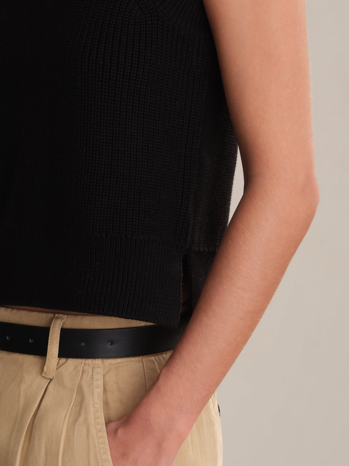 A person wearing a black 100% cotton sleeveless knit top with a crew neck from 525 America and beige pleated pants with a black belt, shown waist up with one hand in their pocket—perfect for effortless layering.