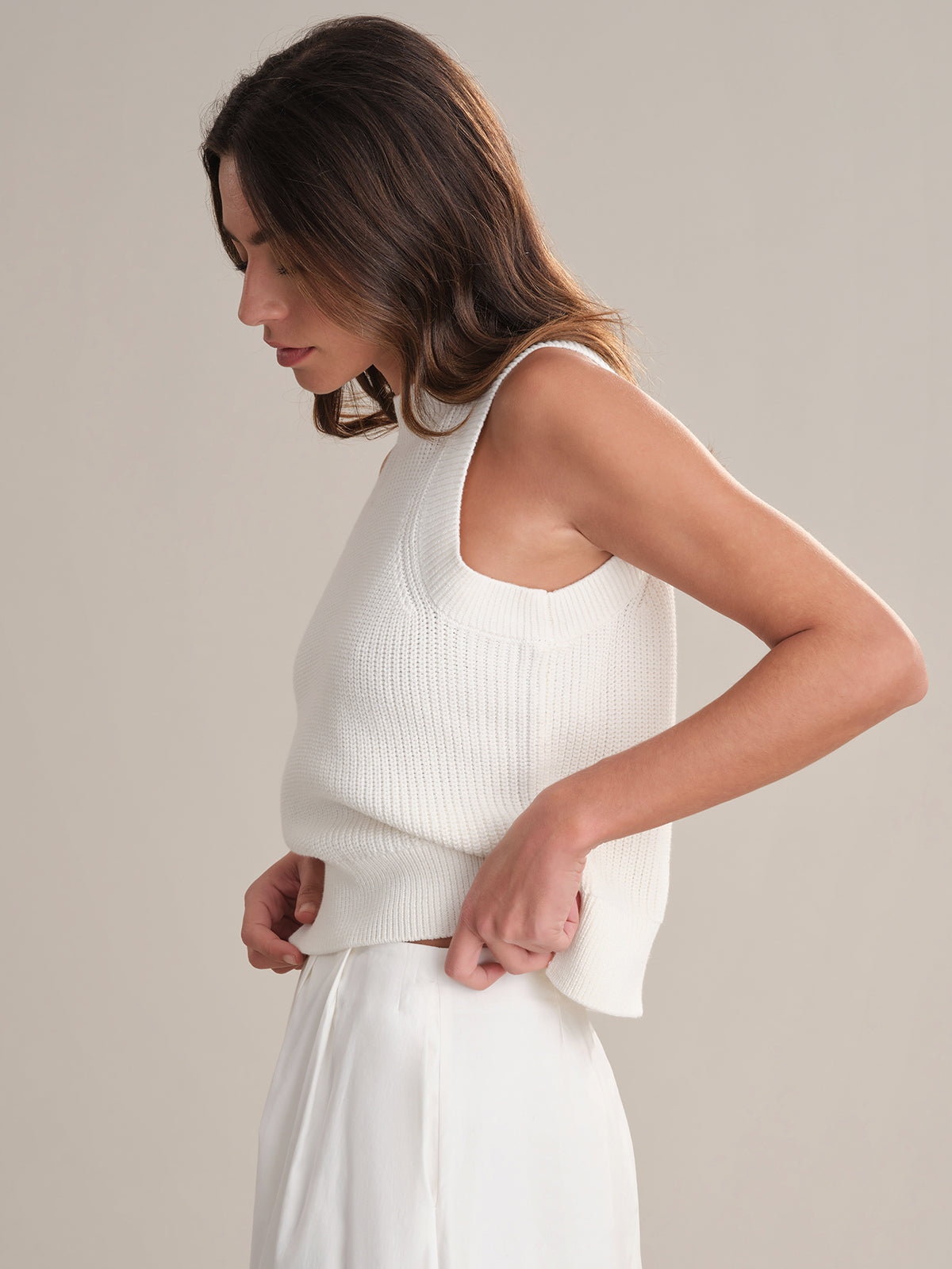 A woman with long brown hair wears a sleeveless white crew neck knit top and skirt from 525 America, standing against a plain light background and looking down as she casually adjusts her top.