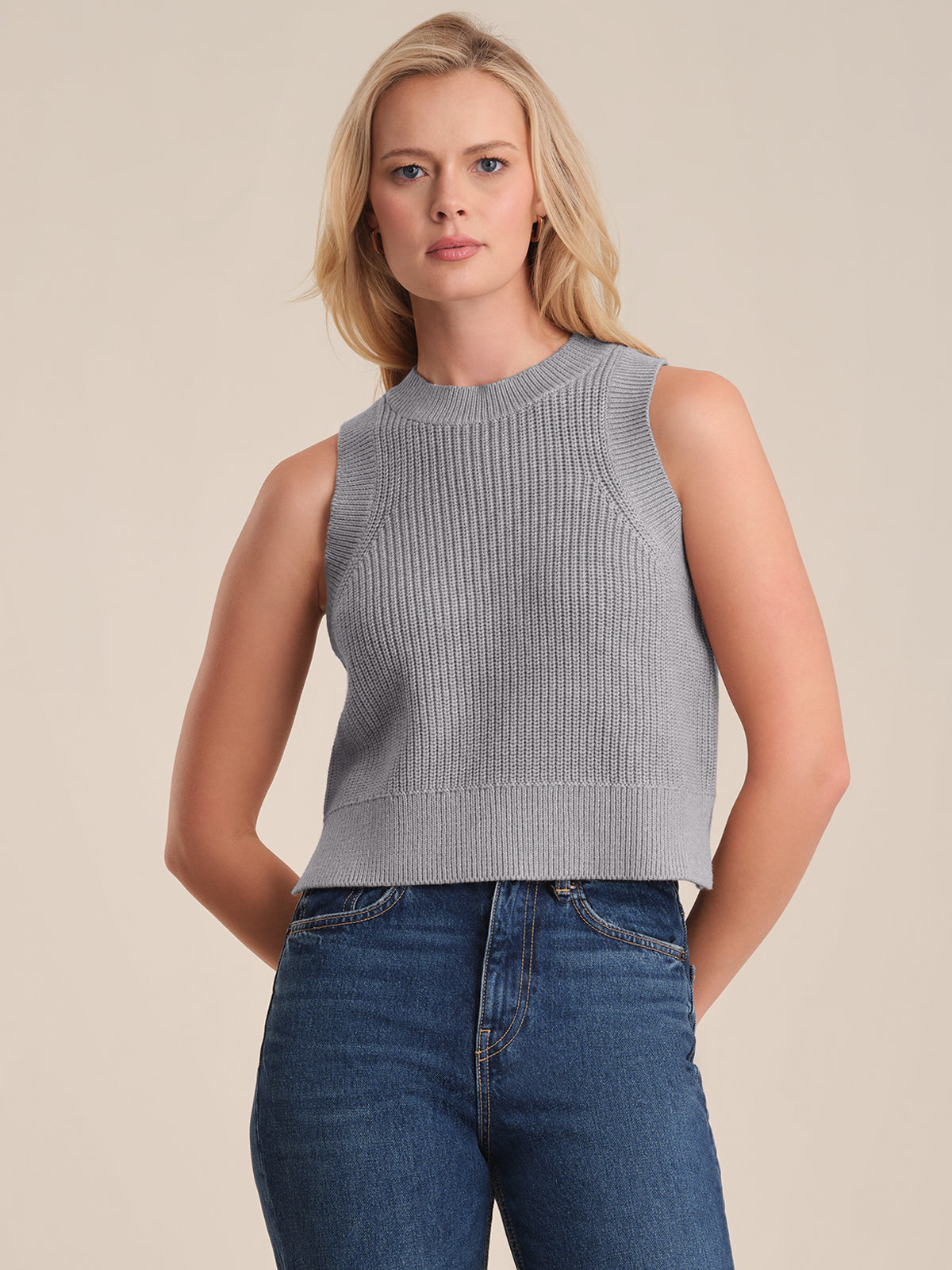 A person with long blonde hair wears a fitted, ribbed gray knit top by 525 America and high-waisted blue jeans, standing against a plain beige background.