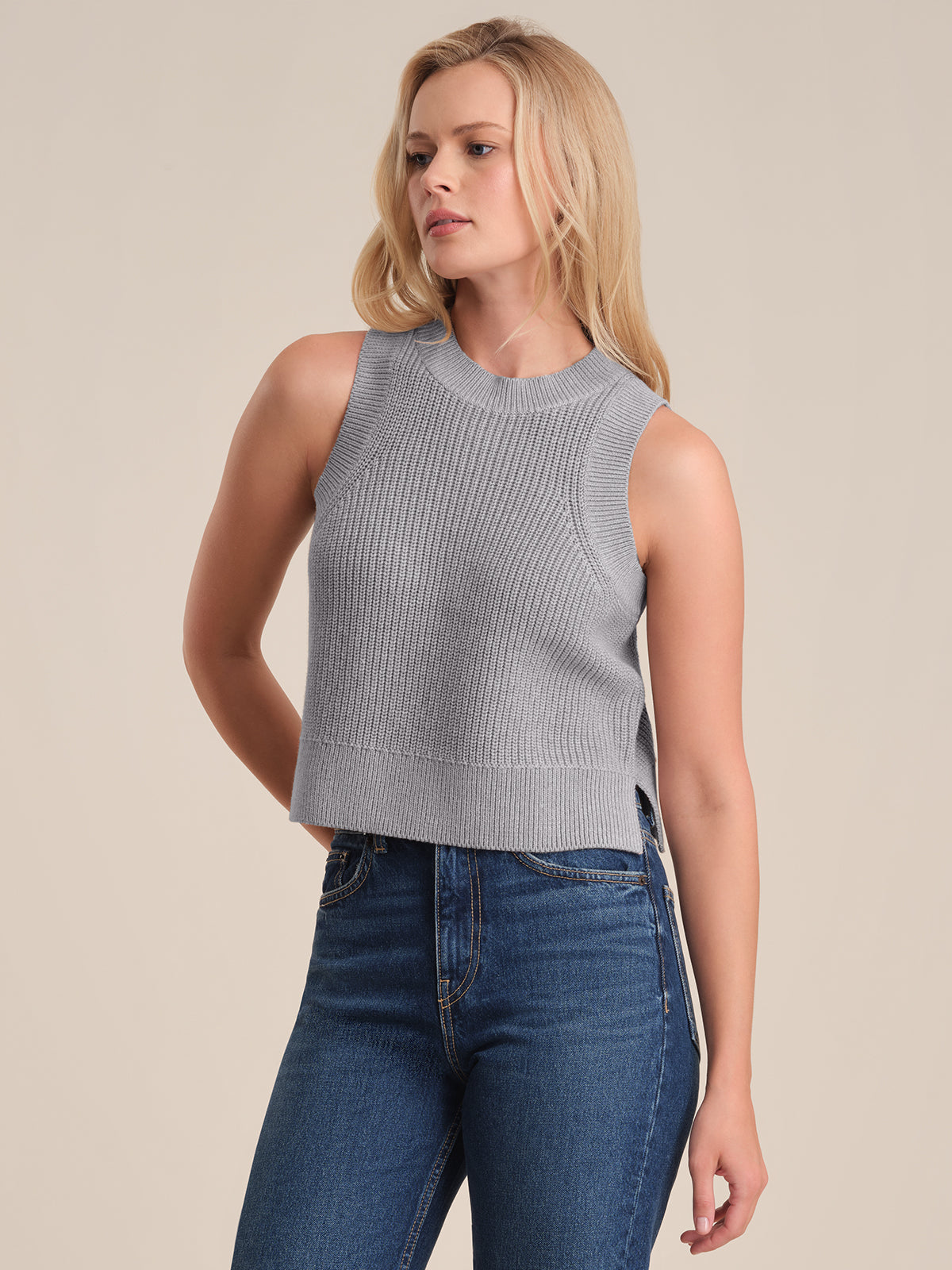 A woman with long blonde hair models a light gray ribbed knit top by 525 America, paired with high-waisted blue jeans. She stands against a beige background, gazing to the side—her look is sophisticated and understated.