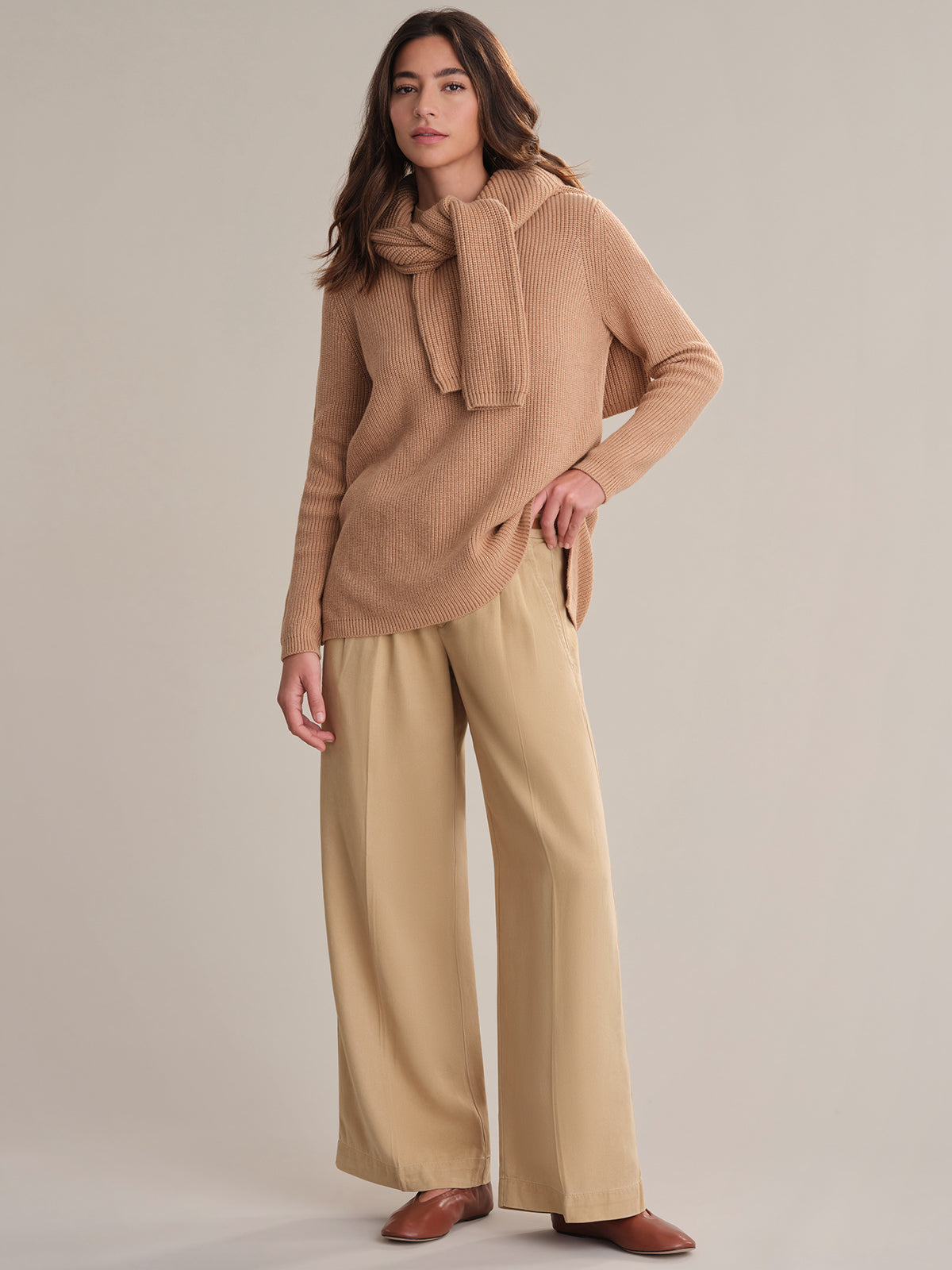 A person stands against a neutral background wearing a relaxed fit beige shaker stitch knit sweater from 525 America, with a matching sweater over their shoulders, wide-leg tan pants, and brown shoes. One hand is in their pocket, the other by their side.