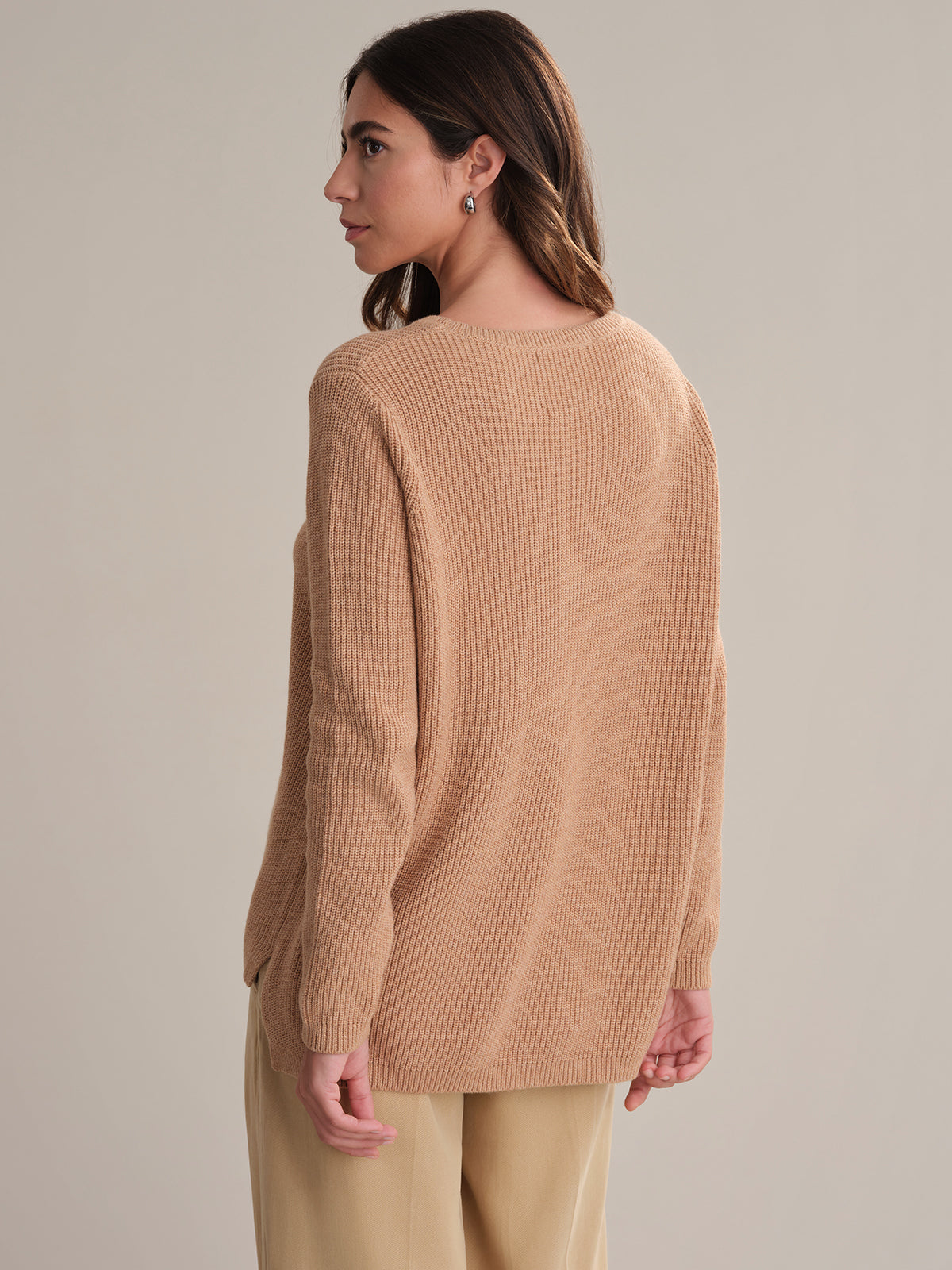 A woman with long brown hair, dressed in a beige 100% cotton ribbed sweater by 525 America and light-colored pants, stands facing away and looking left against a plain light background.