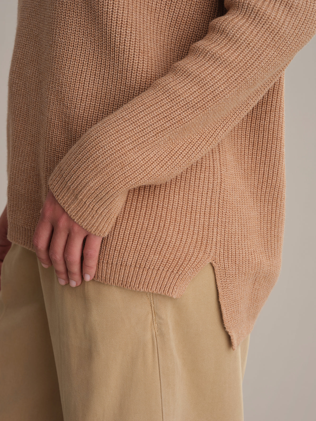 A person wears a 525 America relaxed fit beige knit sweater with ribbed cuffs and hem, paired with light tan pants. Their left arm rests at their side. A neutral background highlights the versatile shaker stitch design.