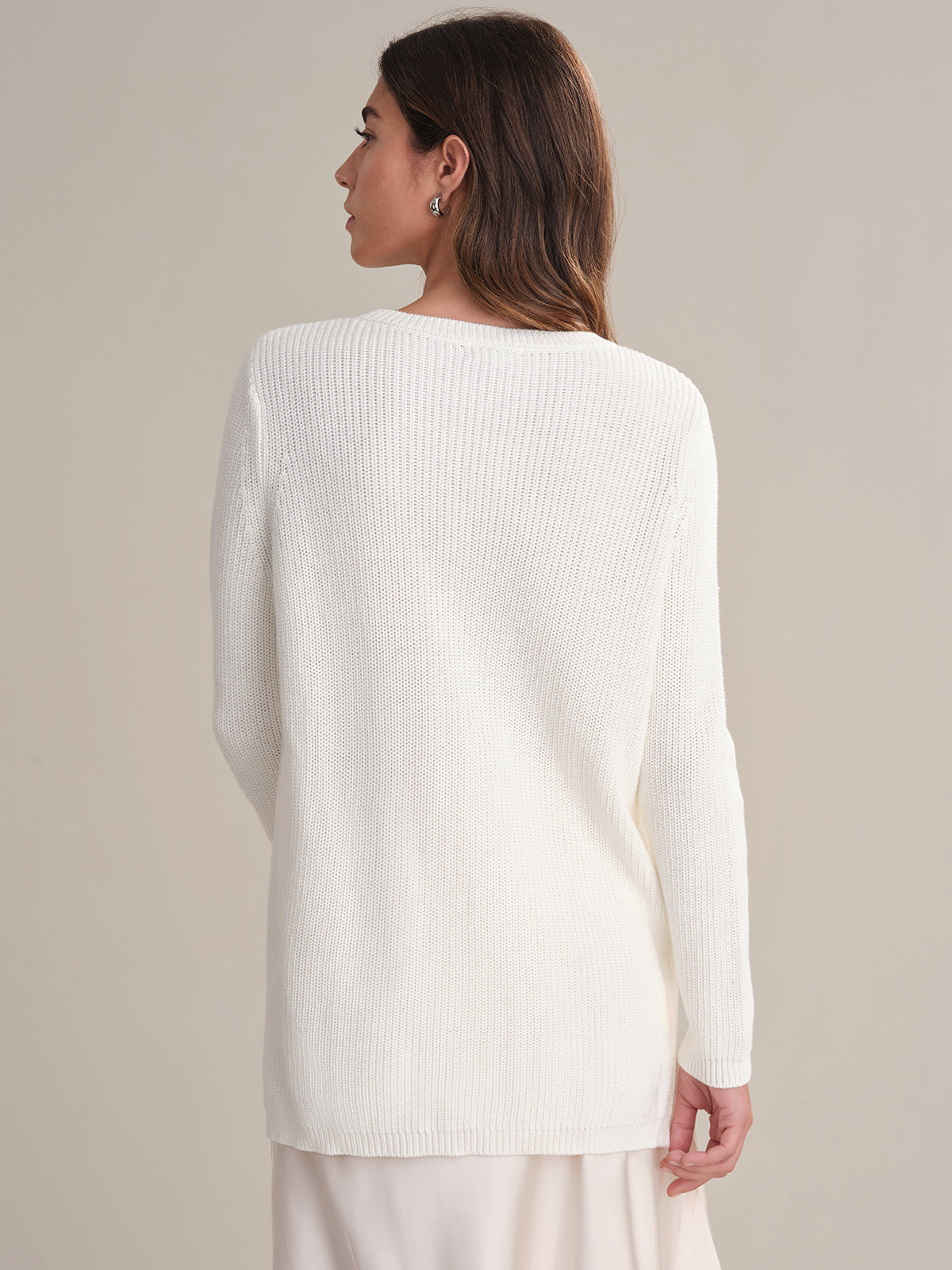 A woman with long brown hair is seen from behind wearing a 525 America white ribbed knit shaker stitch sweater and a light skirt, standing against a simple, neutral background.