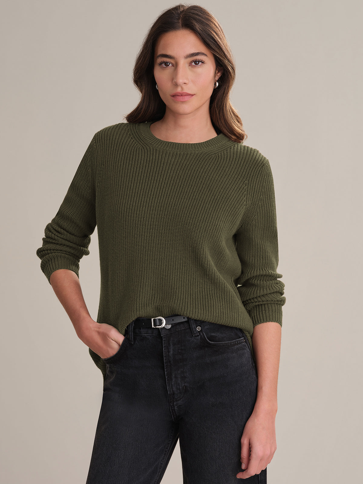 A woman with long brown hair models a 525 America olive green ribbed shaker stitch sweater and dark jeans with a black belt, standing against a plain beige background. She faces the camera with one hand in her pocket and a neutral expression.