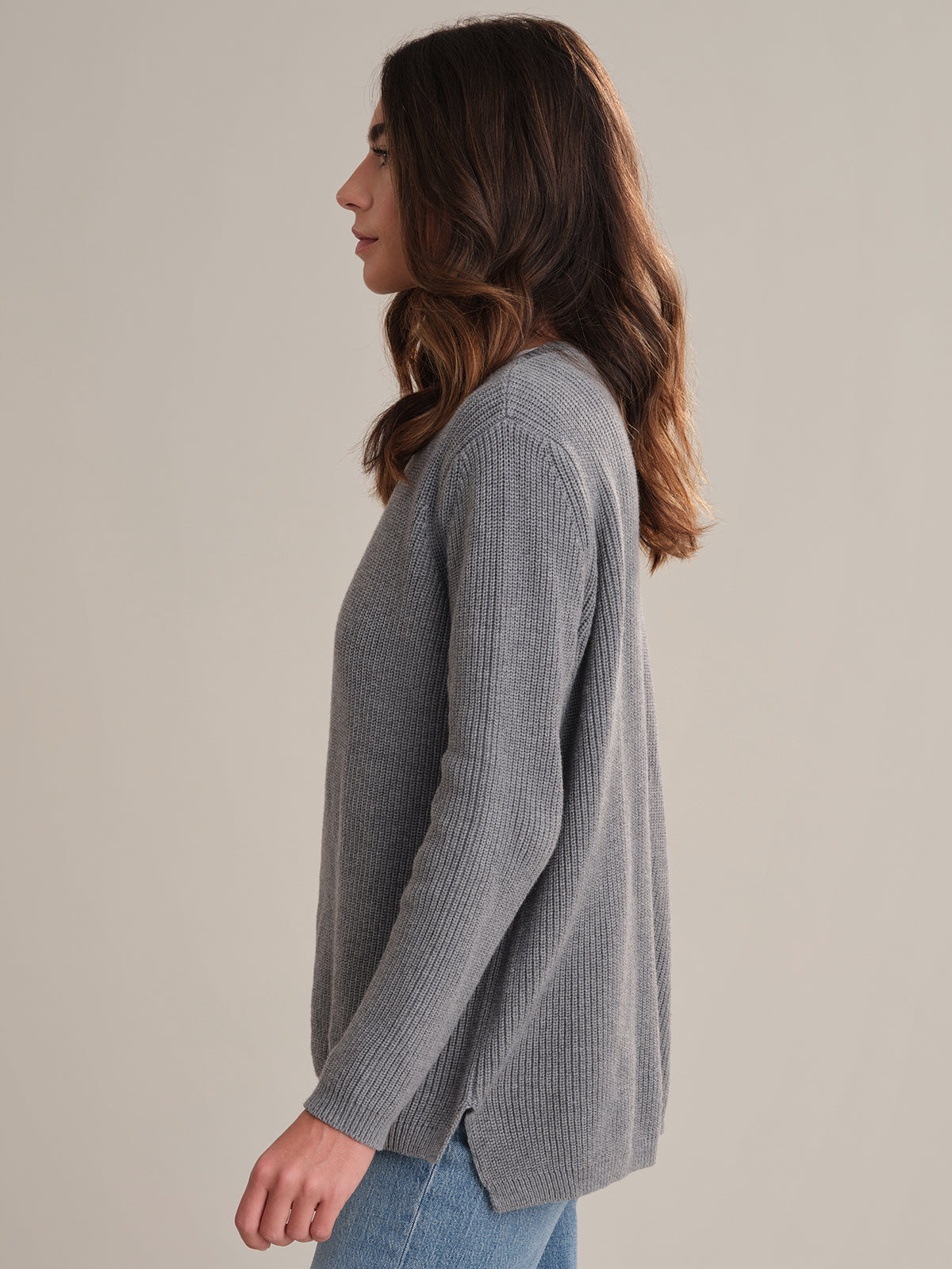A person with long brown hair stands in profile against a plain background, wearing a relaxed-fit, versatile gray shaker stitch sweater by 525 America and blue jeans.