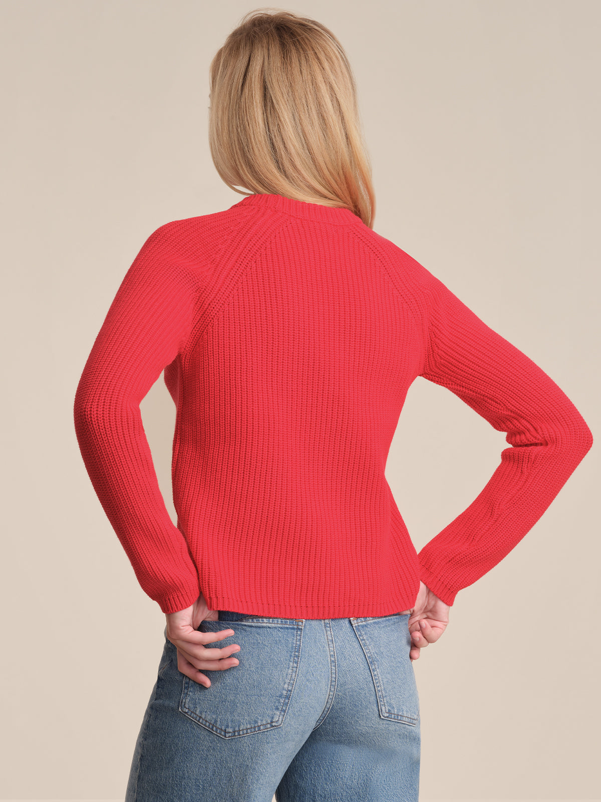 A person with long blonde hair is seen from the back, wearing a 525 America bright red cotton shaker stitch fitted sweater and blue jeans, standing with one hand on their hip against a neutral background.