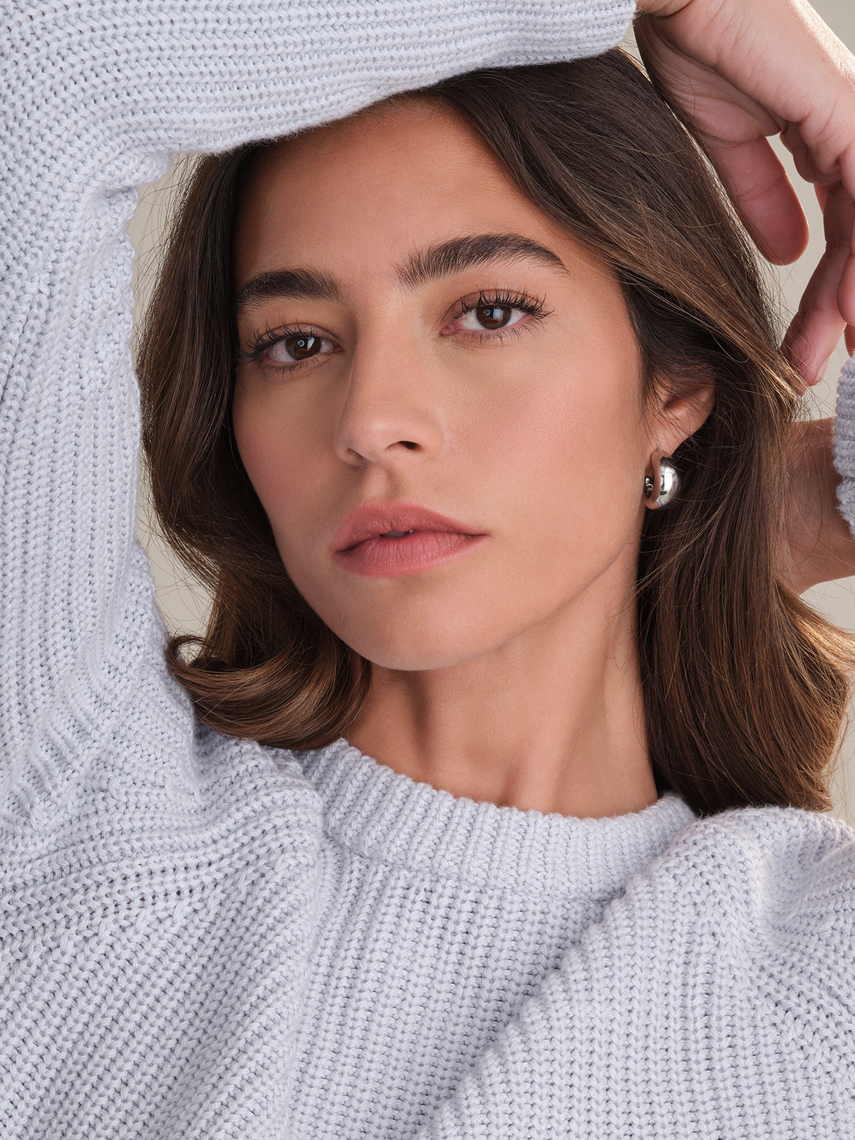 A woman with long brown hair and neutral makeup models a 525 America cotton shaker stitch sweater with raglan sleeves and a fitted silhouette, silver hoop earrings, and poses confidently, looking directly at the camera.