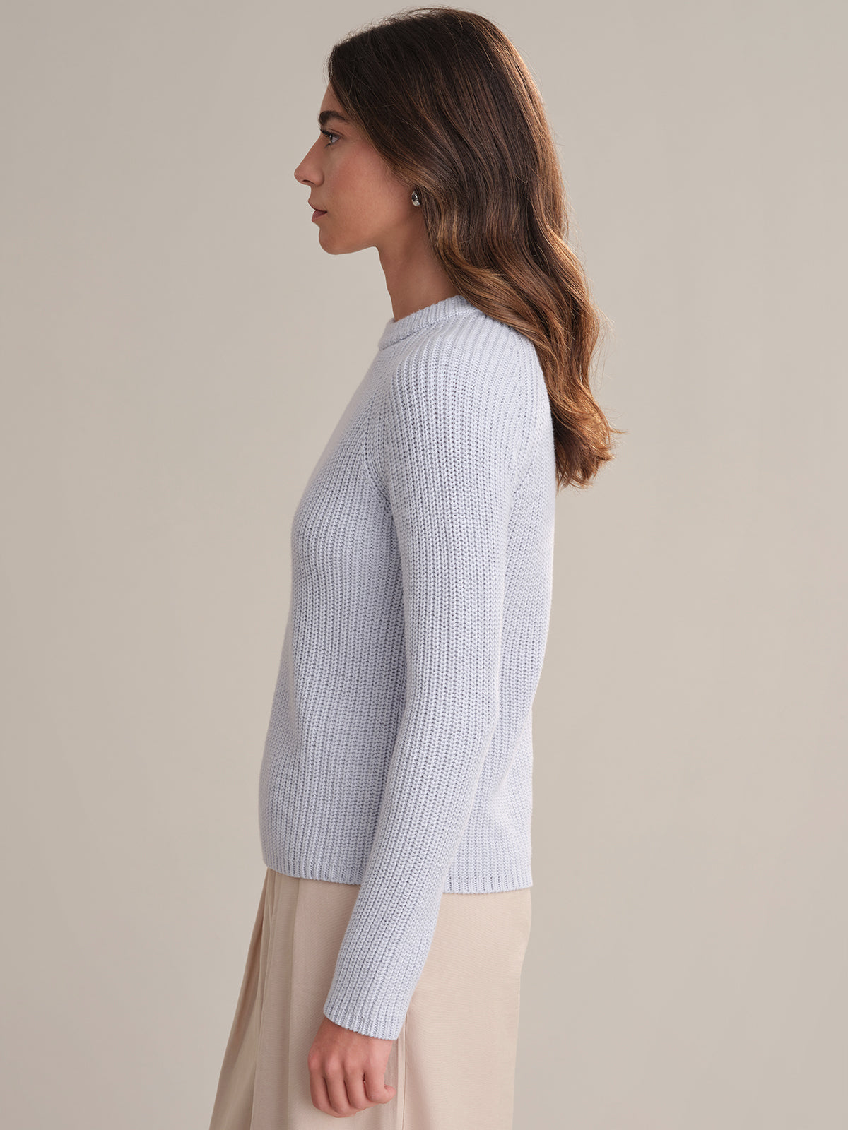 A woman with long brown hair, dressed in a fitted light blue cotton shaker stitch sweater by 525 America and a beige skirt, stands in profile against a plain, light background.