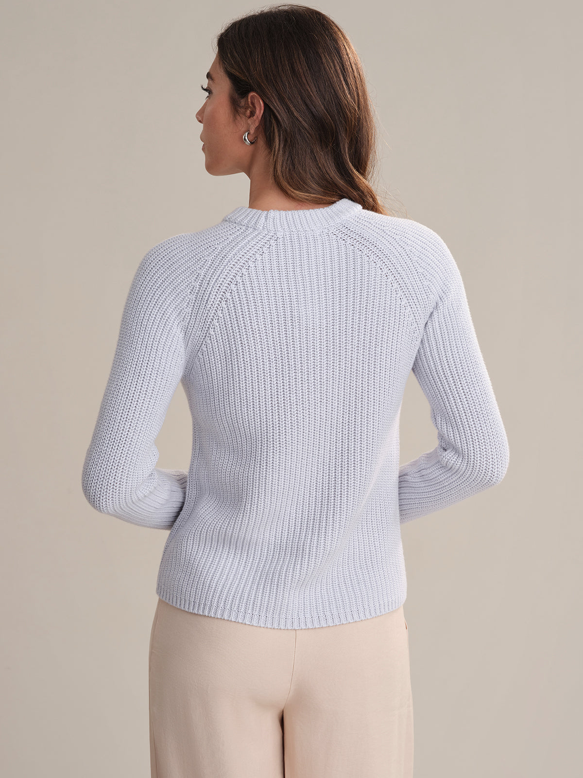 A woman is seen from behind in a fitted, light-colored cotton shaker stitch sweater by 525 America and beige pants, set against a plain neutral background.