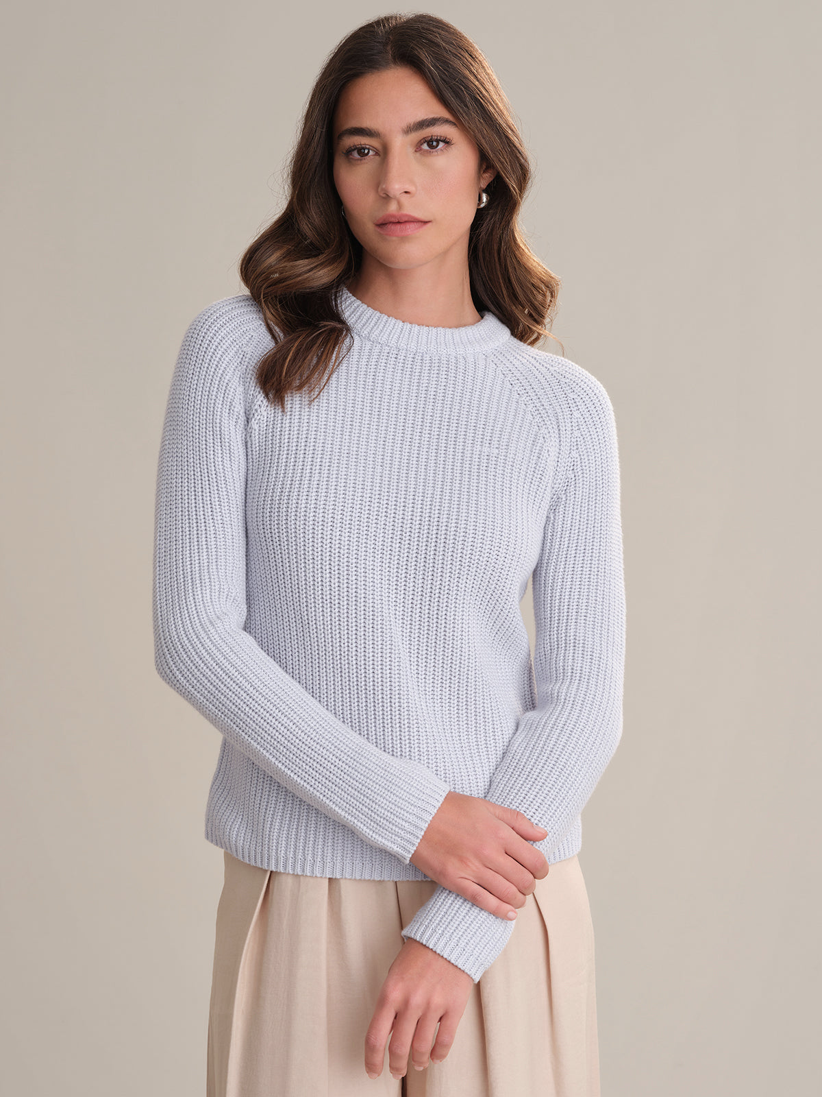 A woman with long brown hair wears a fitted, light gray cotton shaker stitch sweater by 525 America and a beige skirt, standing before a plain light background and looking at the camera with a neutral expression.