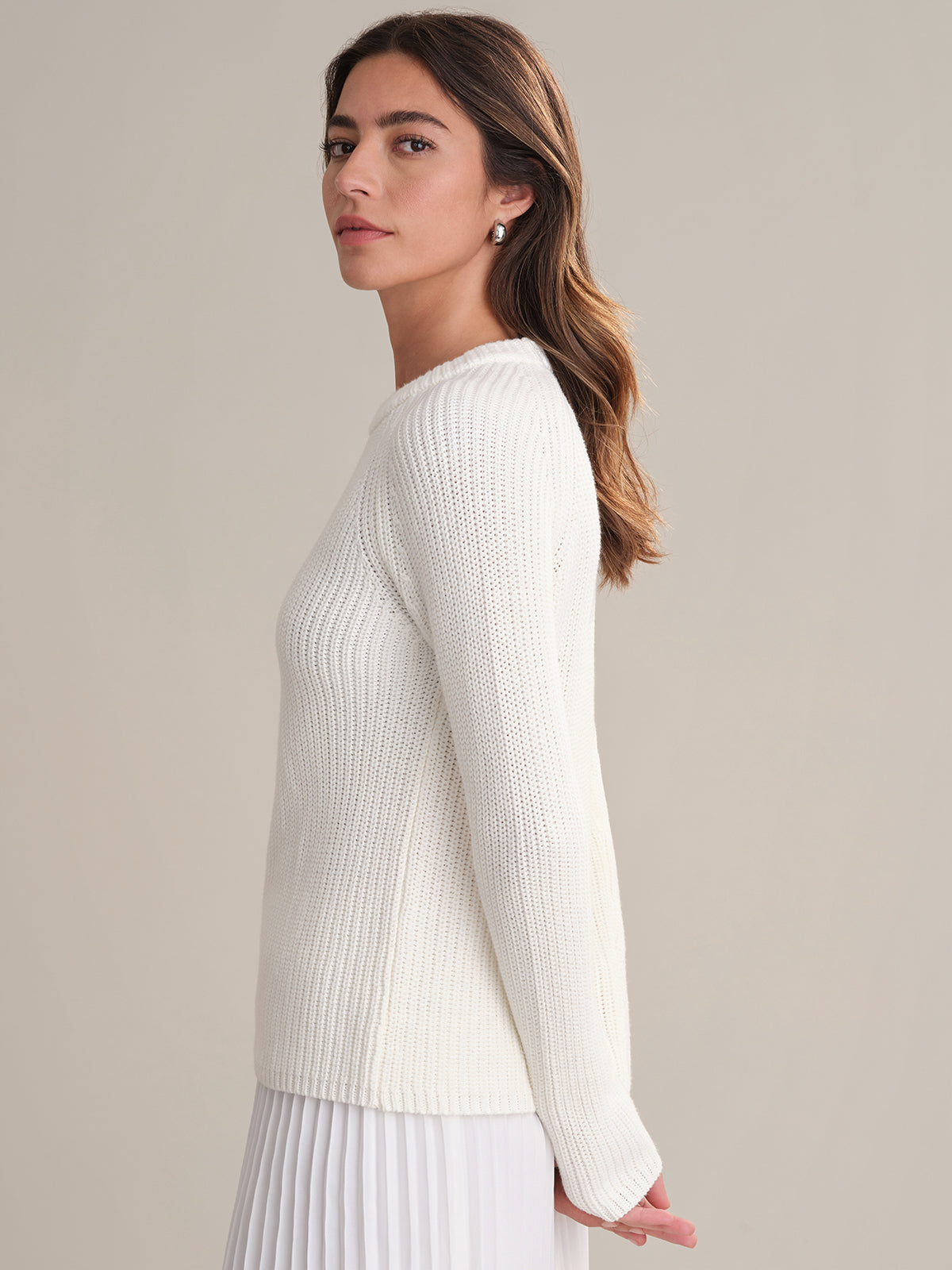 A woman with long brown hair, wearing a 525 America white cotton shaker stitch sweater with raglan sleeves and a white pleated skirt, stands in profile against a plain light beige background.