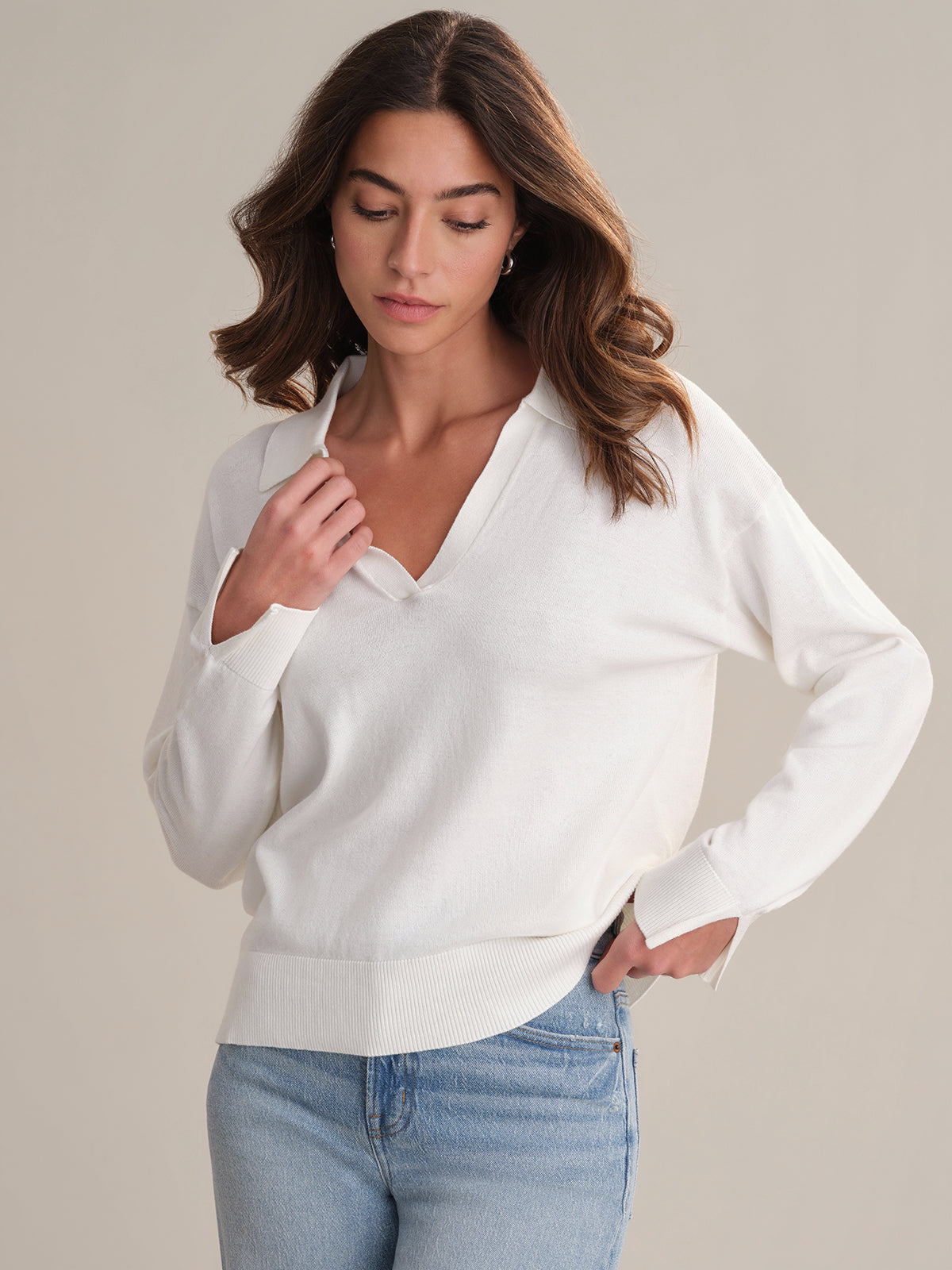 A woman models a 525 America white lightweight Johnny collar pullover with light blue jeans, standing against a plain beige background. She looks down, touching her collar with one hand and keeping the other in her pocket.