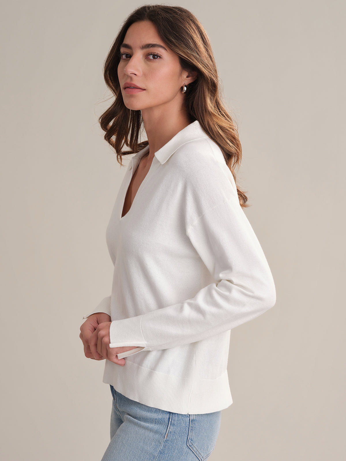 A woman with long brown hair models a white, breathable Johnny collar sweater by 525 America and light blue jeans, standing in profile against a plain beige background.