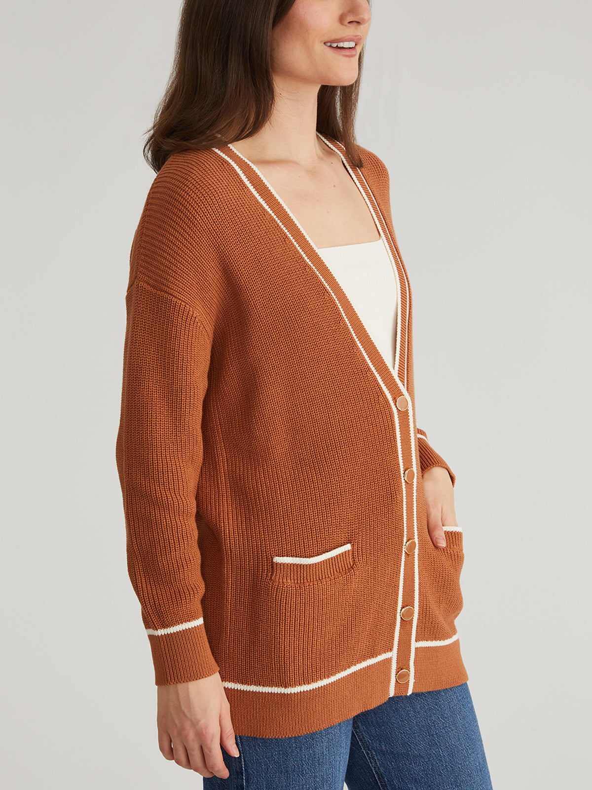A person models a rust-colored 525 America grandpa-style cardigan with white trim and buttons over a white top and blue jeans. Their head is mostly cropped out as they pose casually with one hand in their pocket against a plain gray background.