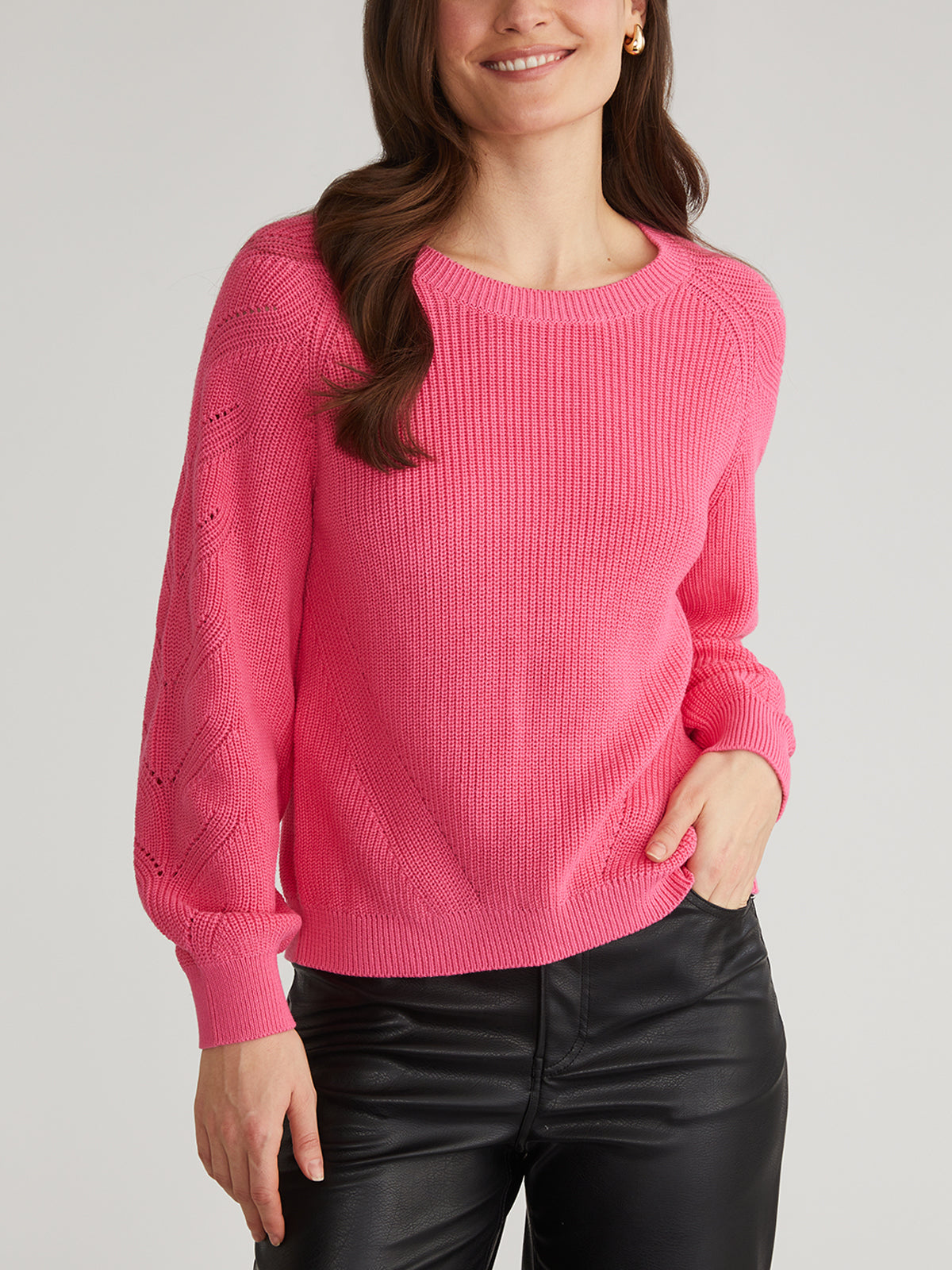 A person is wearing a vibrant pink sweater by 525 America, featuring textured Raglan Pointelle Sleeves, paired with black leather pants. Their hair is long and wavy as they stand against a plain background.