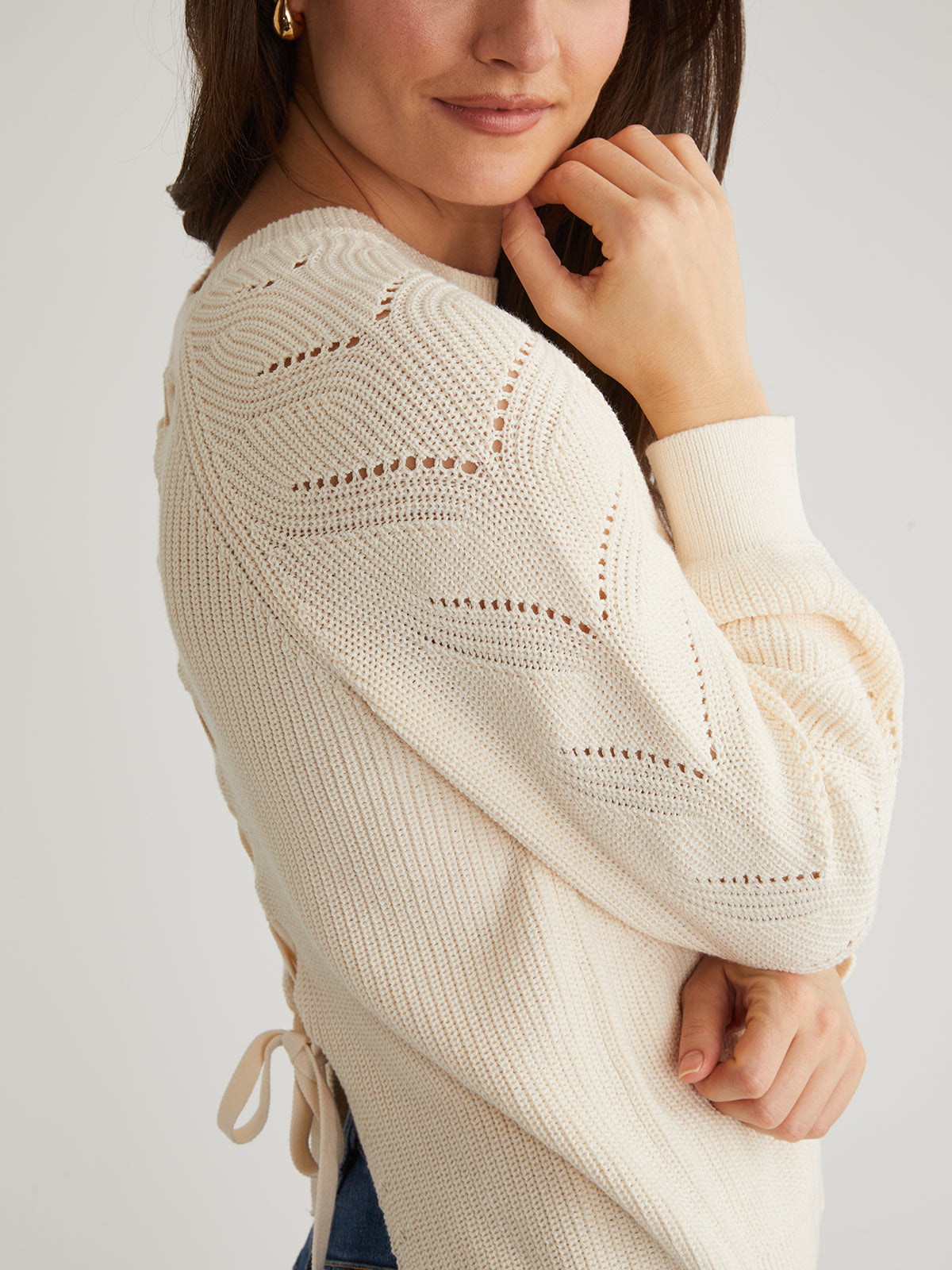 A person is in a cream 525 America knitted sweater with raglan pointelle sleeves and decorative patterns, touching their face with one hand and hair tucked behind the ear. The plain neutral background enhances the cozy feel of the 100% cotton garment.