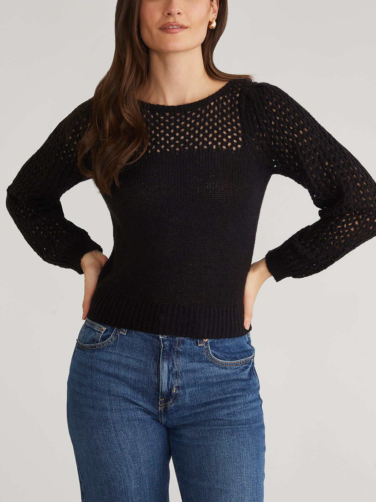 The individual wears a 525 America black knitted sweater with breathable mesh on the upper section and sleeves, adding a modern flair. Their ensemble is complemented by blue jeans. Long brown hair cascades down as they confidently place their hands on their hips against a plain gray background.