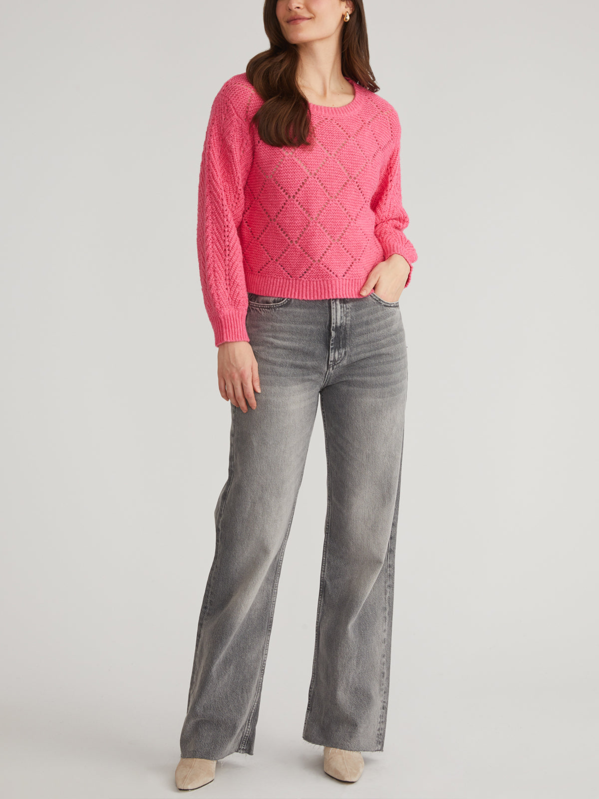 A woman wears a 525 America pink Pointelle Pullover with balloon sleeves and gray wide-leg jeans, one hand in her pocket, hair cascading down. Light-colored shoes complete the look against a neutral background.