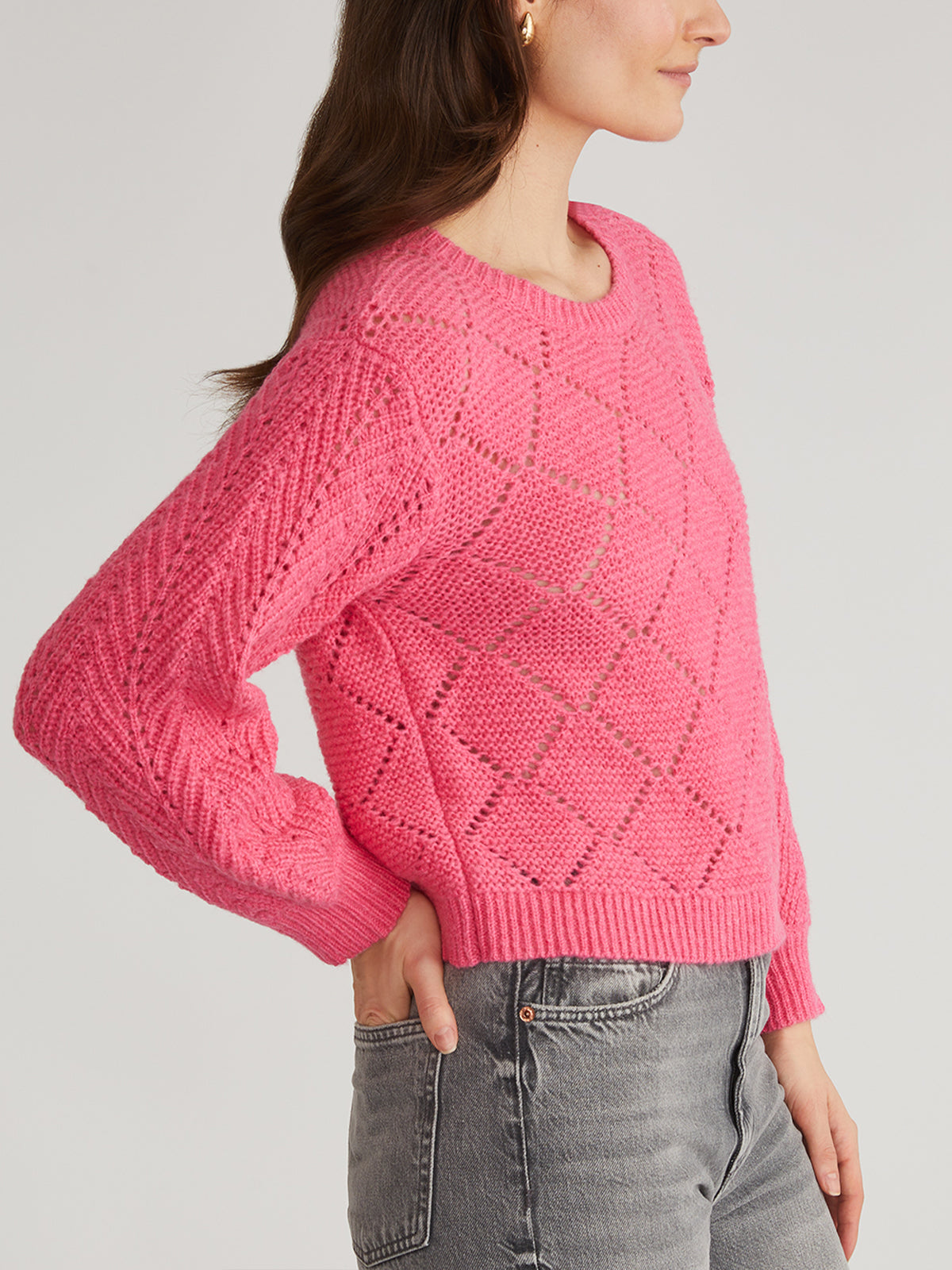 A person in a pink 525 America pointelle pullover, featuring diamond patterns and balloon sleeves, stands in profile. They pair it with high-waisted blue jeans, hand confidently on hip. The plain light gray background accentuates their stylish look.