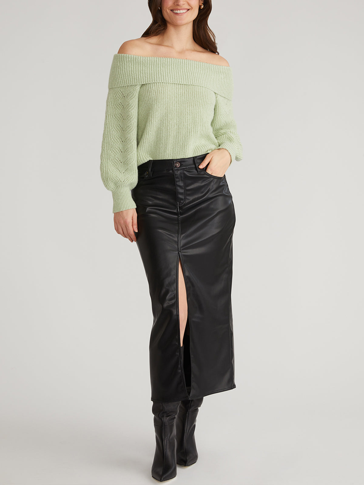 Someone is wearing a 525 America light green off-the-shoulder knit sweater with a relaxed pullover feel, paired with a black leather midi skirt featuring a front slit. Their long brown hair flows down as they stand against a white background, finishing the look with black boots for casual outings.