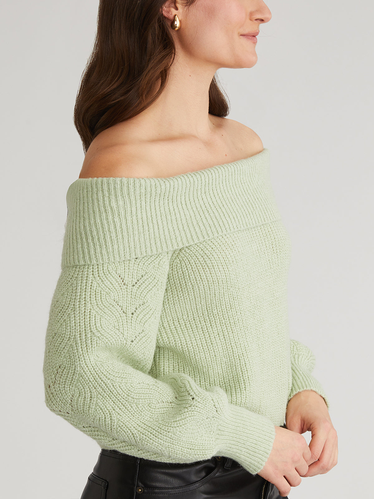 Dressed for a casual outing, the person wears a 525 America light green off-the-shoulder knit sweater featuring cable-patterned sleeves, paired stylishly with black leather pants. The side profile highlights long hair and small hoop earrings, with arms crossed elegantly.