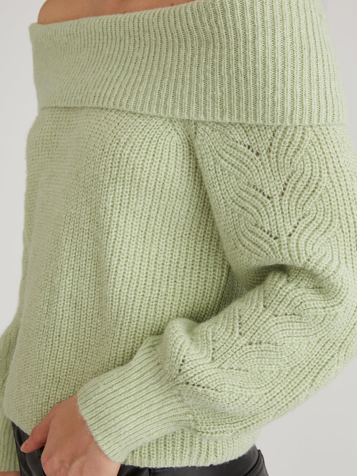 The individual wears a light green off-the-shoulder sweater from 525 America, ideal for casual outings. The relaxed pullover showcases cable knit patterns on the sleeves and is paired stylishly with black pants to accentuate its design and texture.