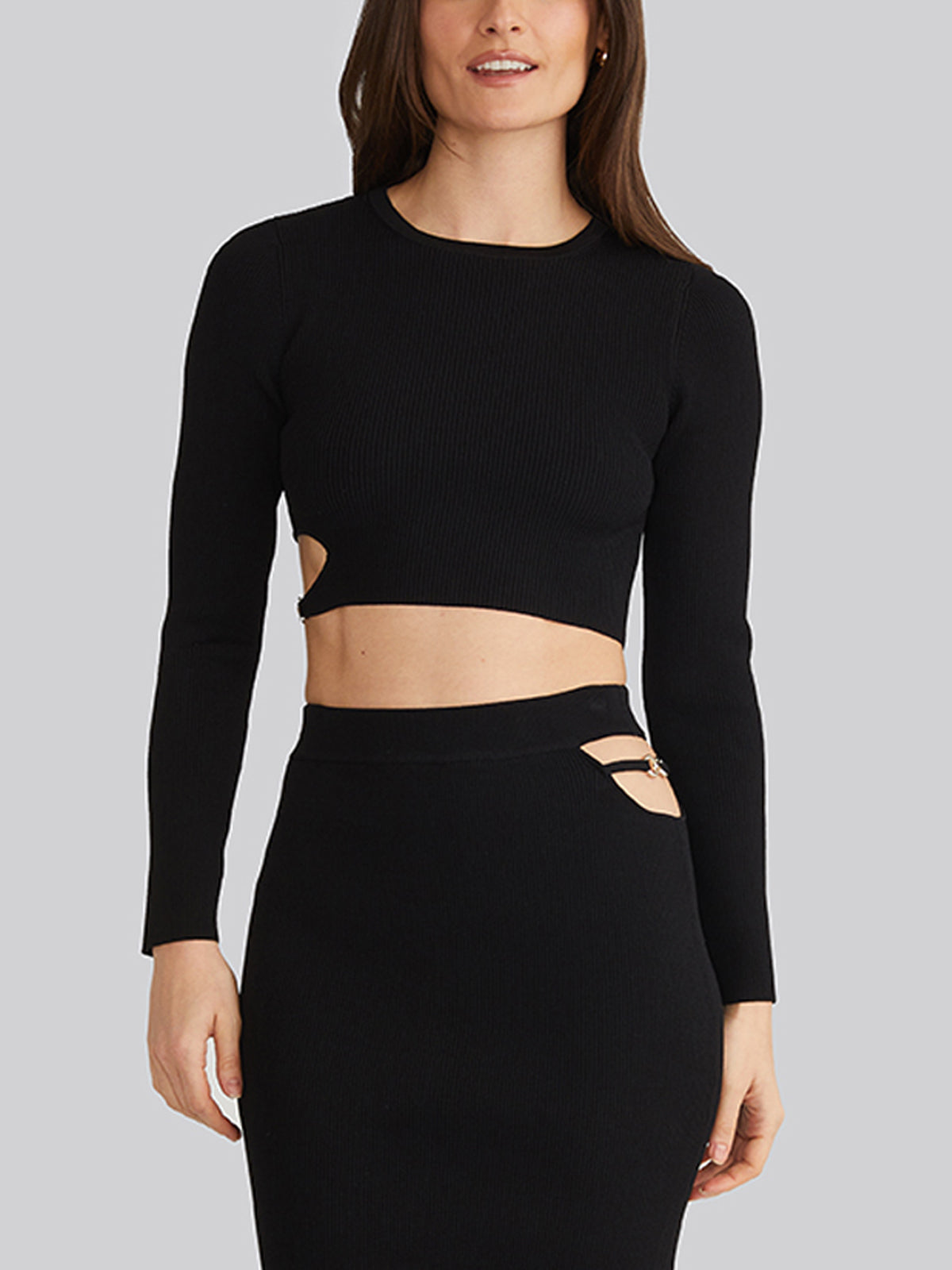 A person models the 525 America Alexia Cropped Buckle Cutout Top, showcasing daring side cutouts, paired with a matching high-waisted skirt featuring its own side cut-out. The ribbed fabric beautifully contrasts with bold buckle details against a gray backdrop.
