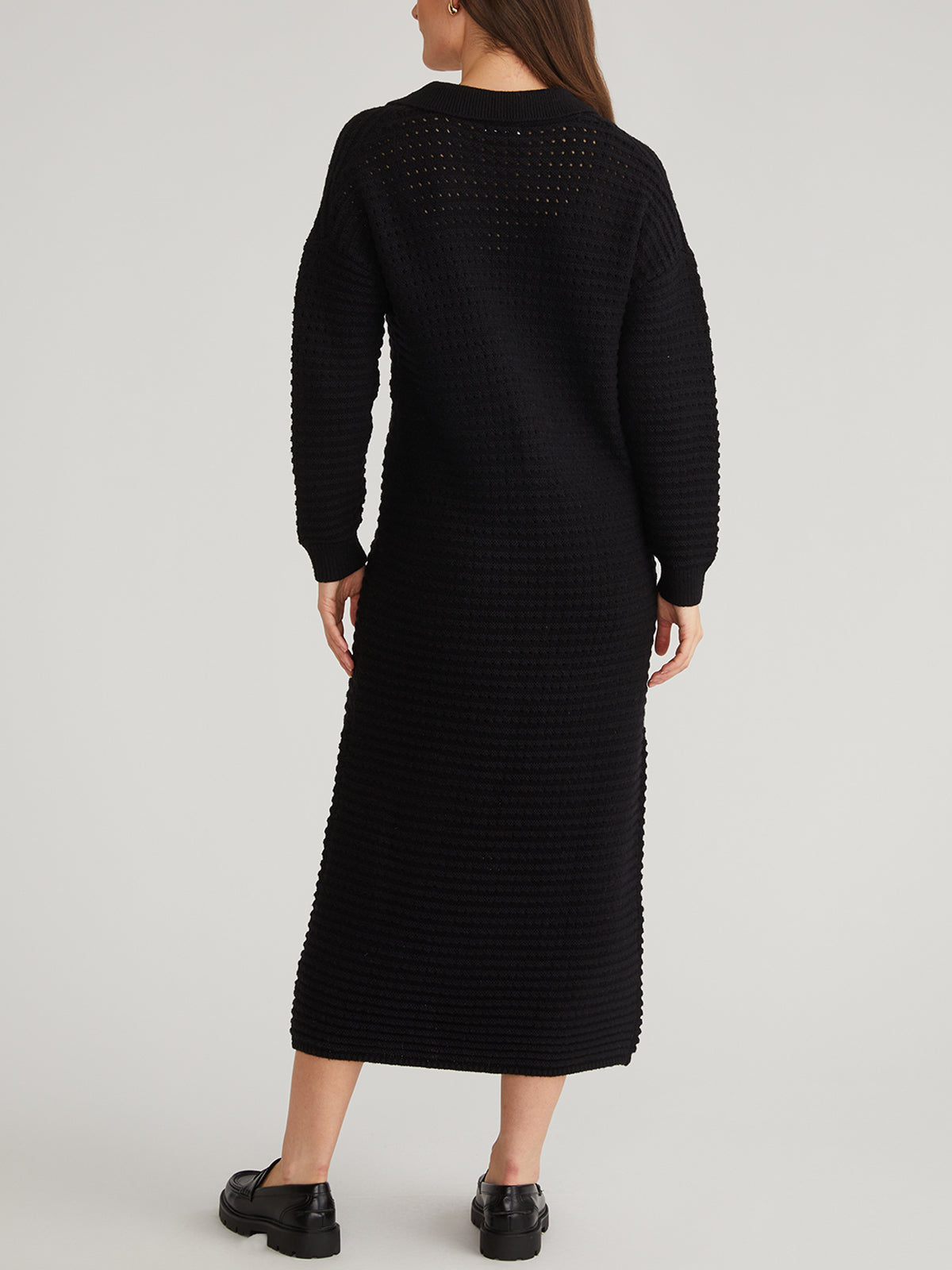 Facing away from the camera against a light background, a person wears an elegant black knit dress with pointelle stitching by 525 America, expertly crafted from heavyweight stretch knit. Their hands rest by their sides, paired with black shoes.