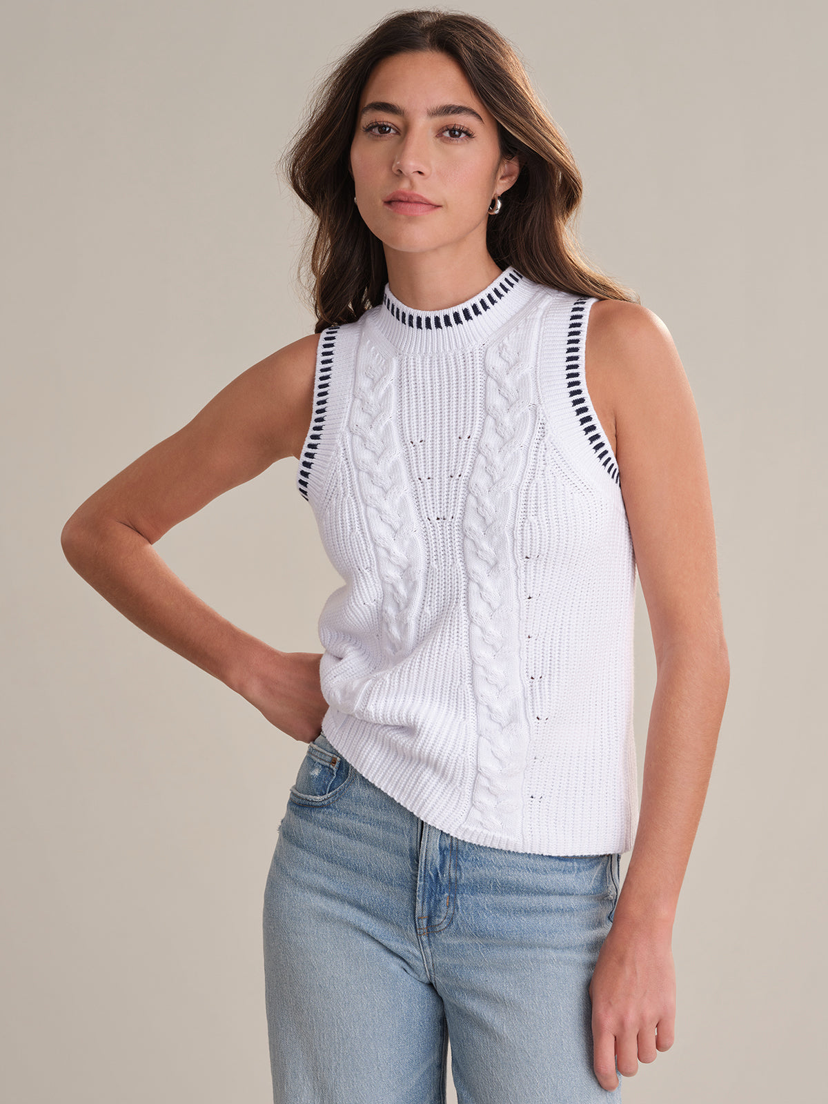 A woman with long brown hair wears a 525 America breathable cotton sweater-tank with double cable stitch, black striped trim, and light blue jeans. She stands against a plain beige background, one hand on her hip.